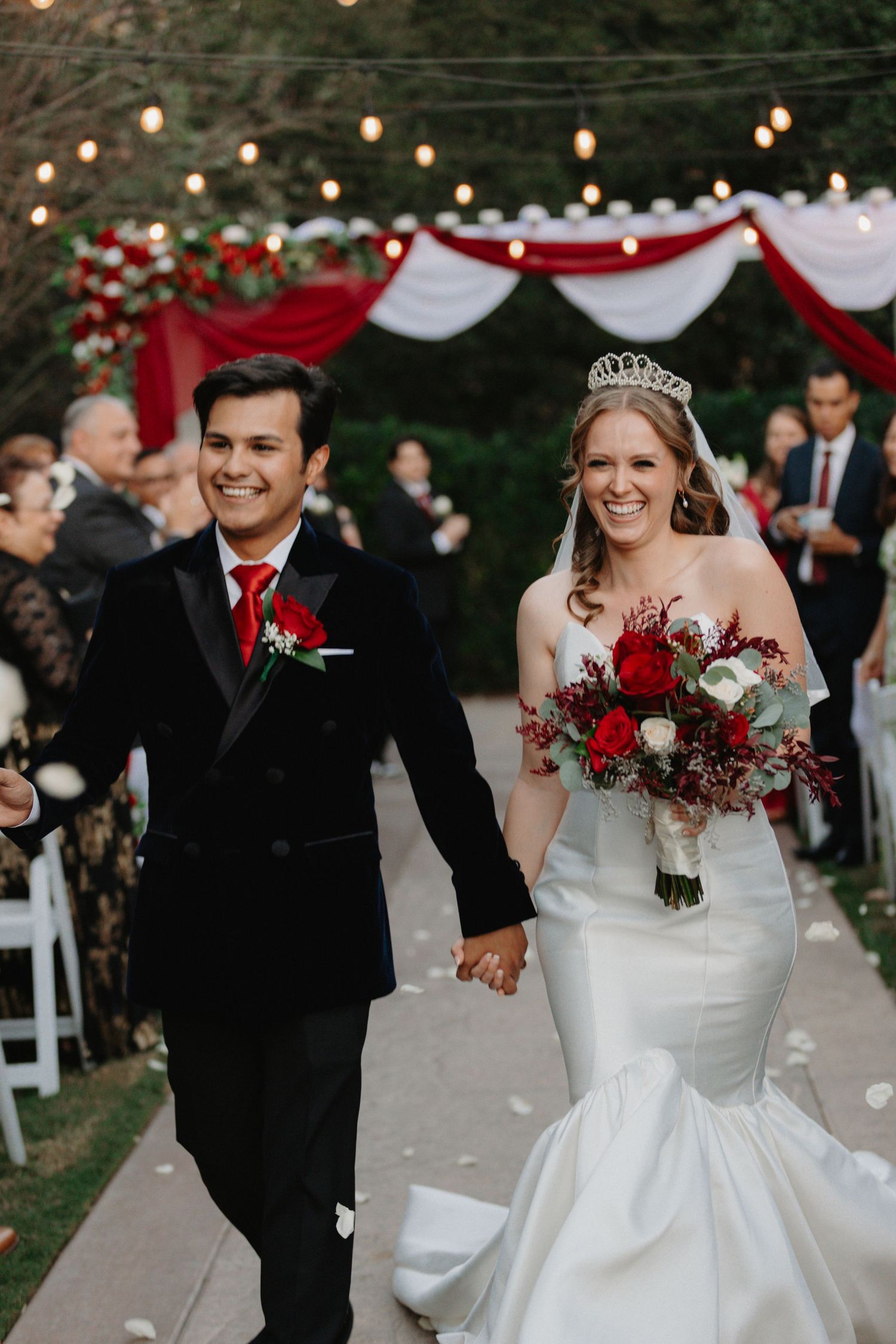 Smiling newlyweds walk down outdoor aisle, bride in tiara holding red bouquet, groom in black tuxedo with red tie.