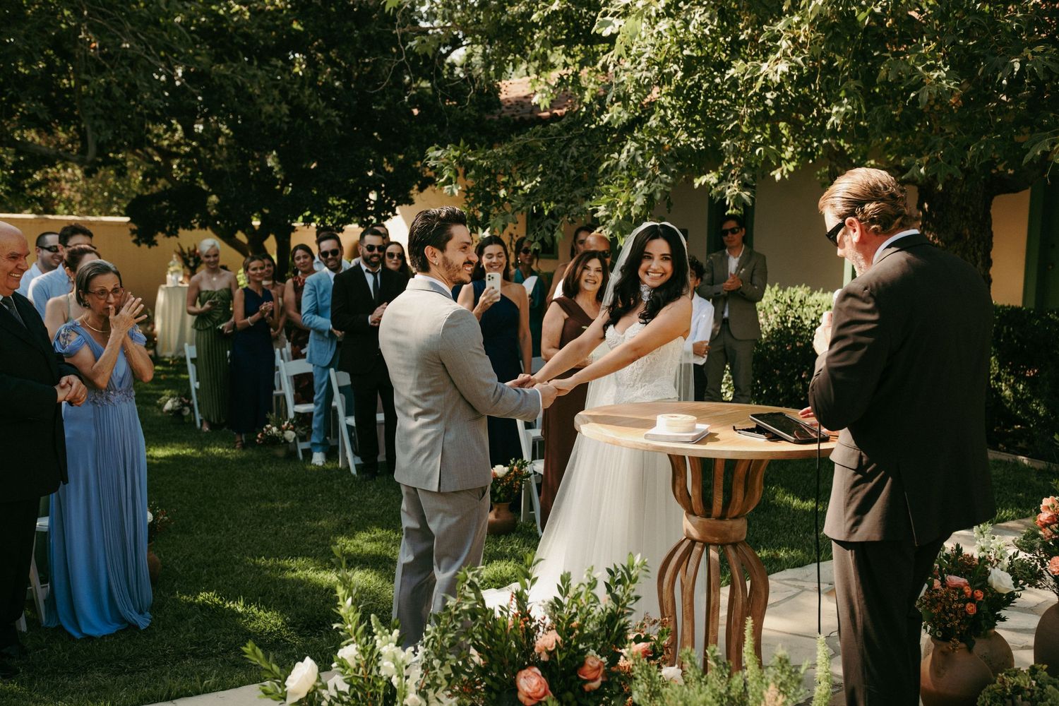 Outdoor wedding ceremony with couple exchanging vows at altar, guests seated on lawn, surrounded by lush trees and floral arrangements.