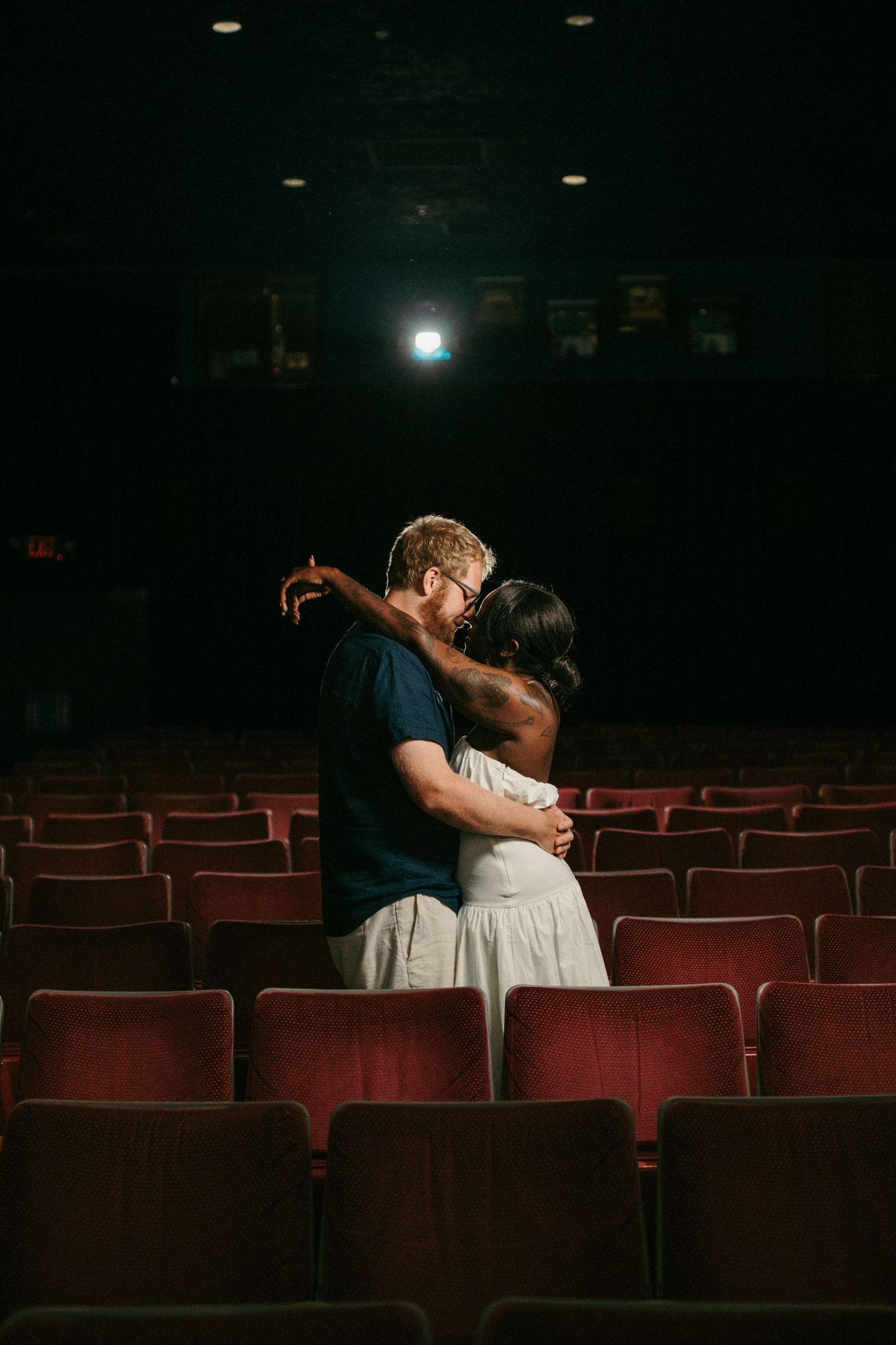 Couple sharing a romantic kiss in an empty red-seated theater with dramatic stage lighting.