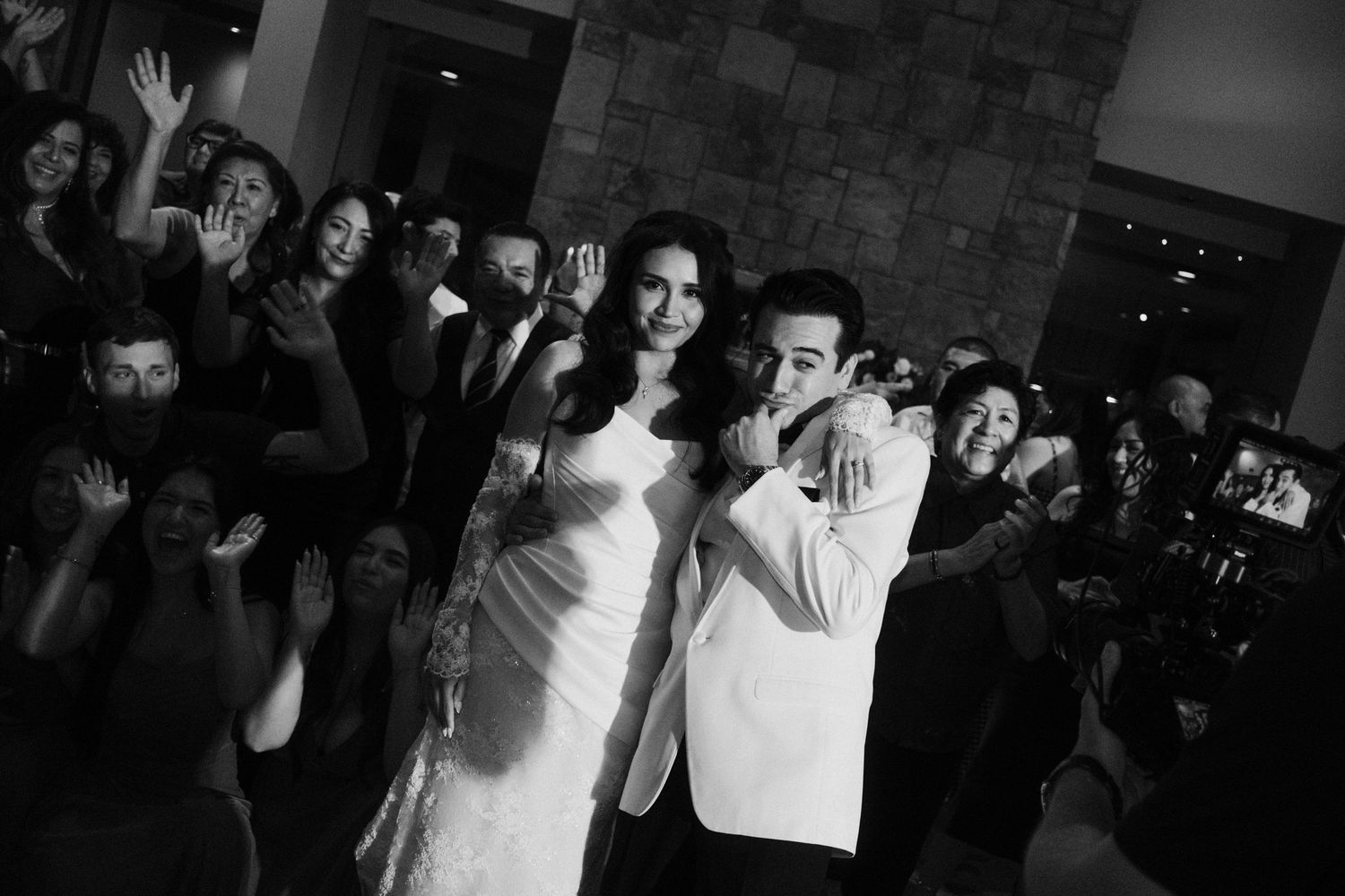 Black and white photo of a bride and groom smiling together surrounded by cheering guests at their wedding reception.