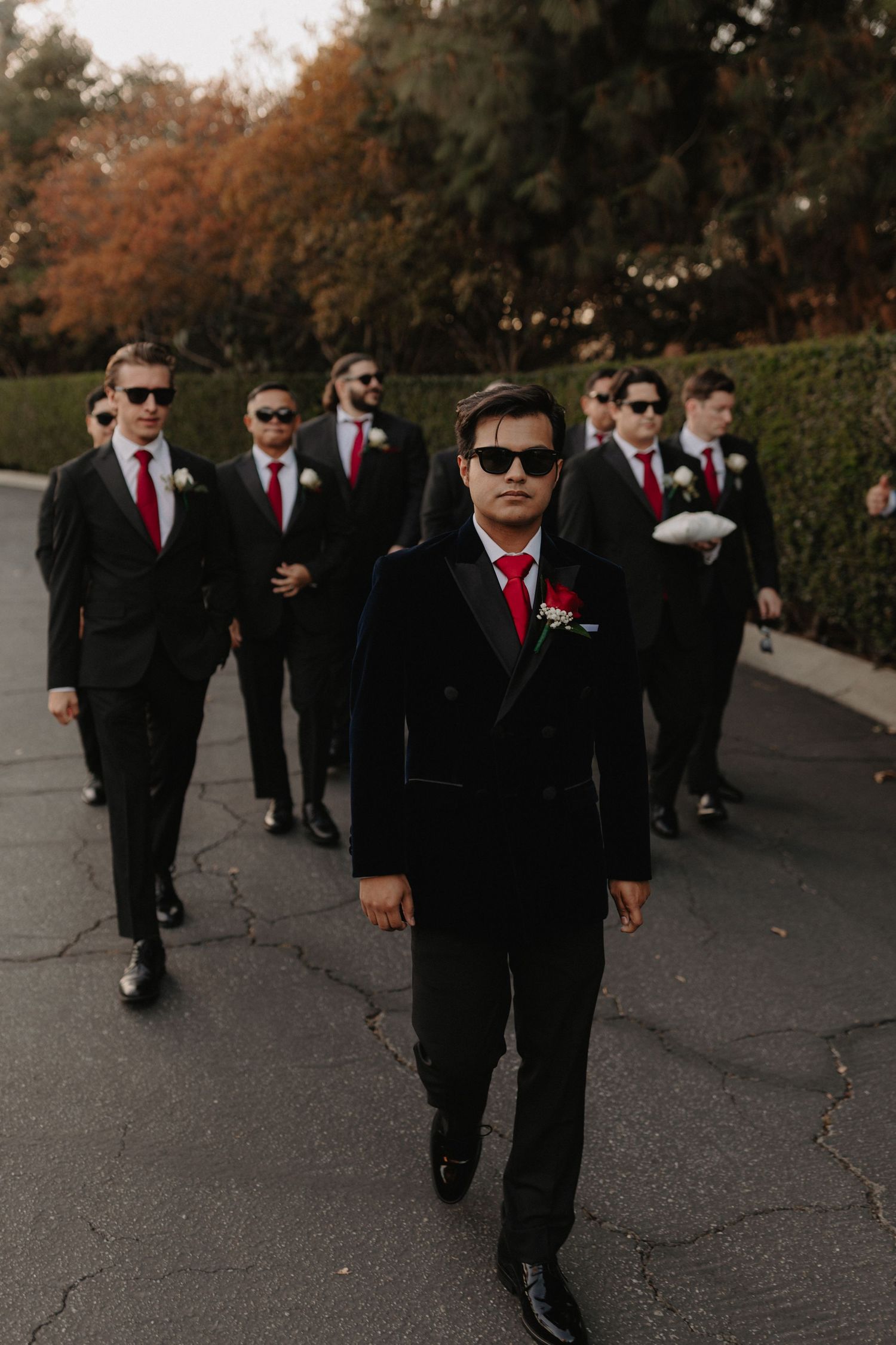 Groom leads groomsmen in black suits with red ties and sunglasses walking outdoors at a wedding.