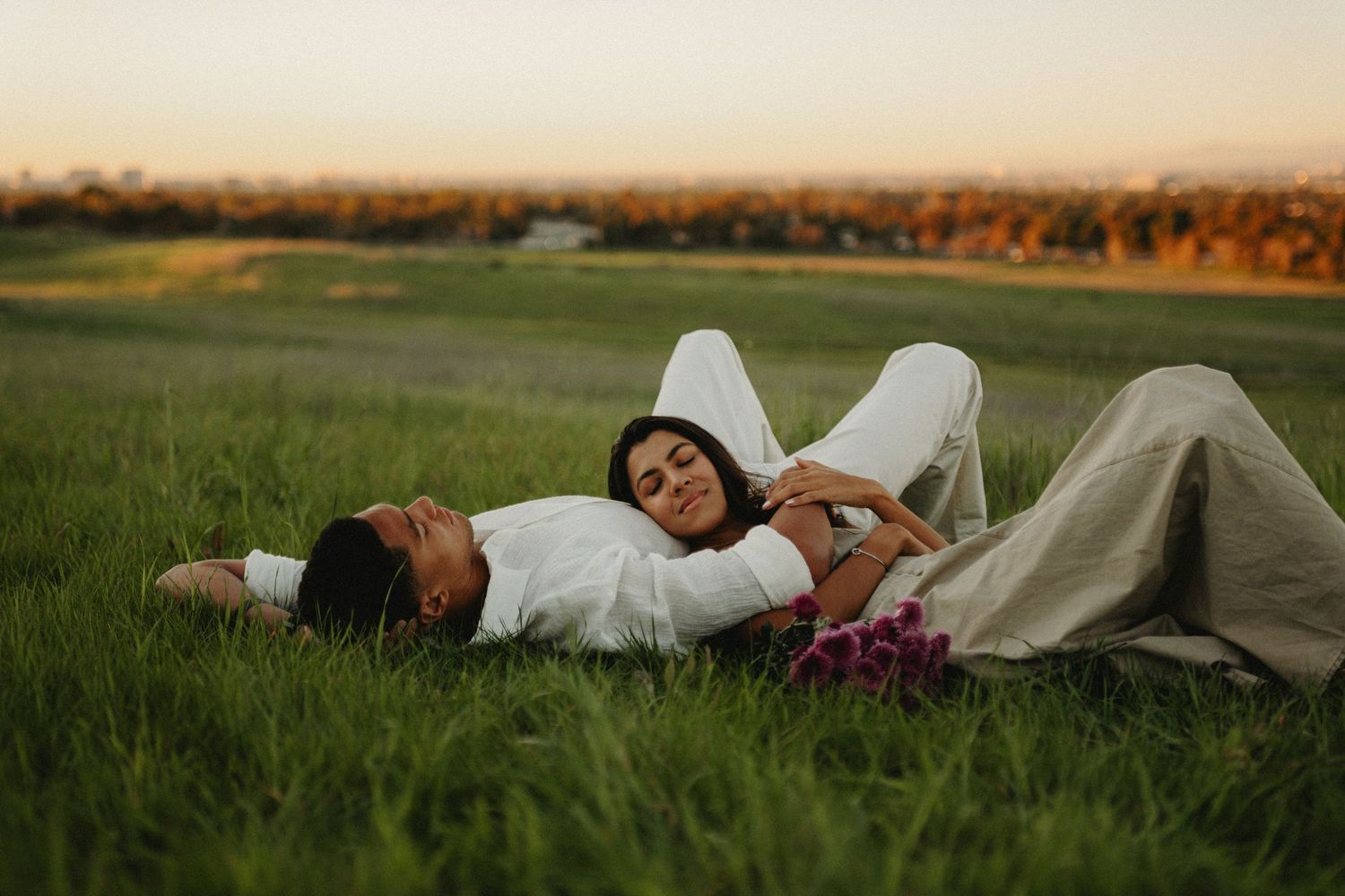 Couple lying together in a lush green field at sunset, woman in white resting on mans chest.