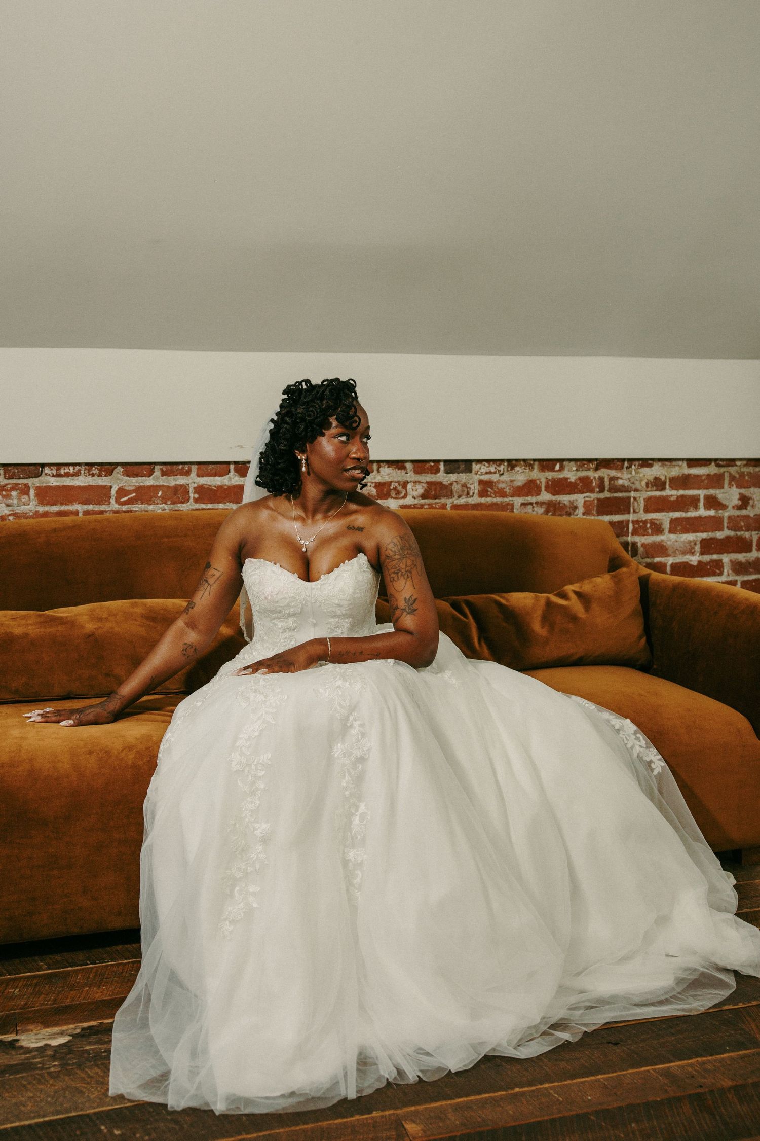 Bride in strapless white ballgown with lace bodice sits gracefully on brown velvet sofa near exposed brick wall.