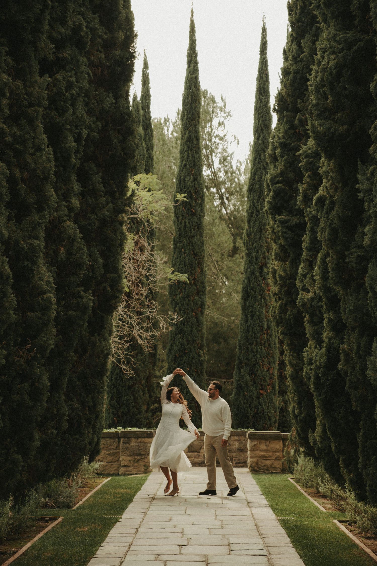 Couple dancing on stone pathway lined with tall cypress trees in a romantic garden setting.