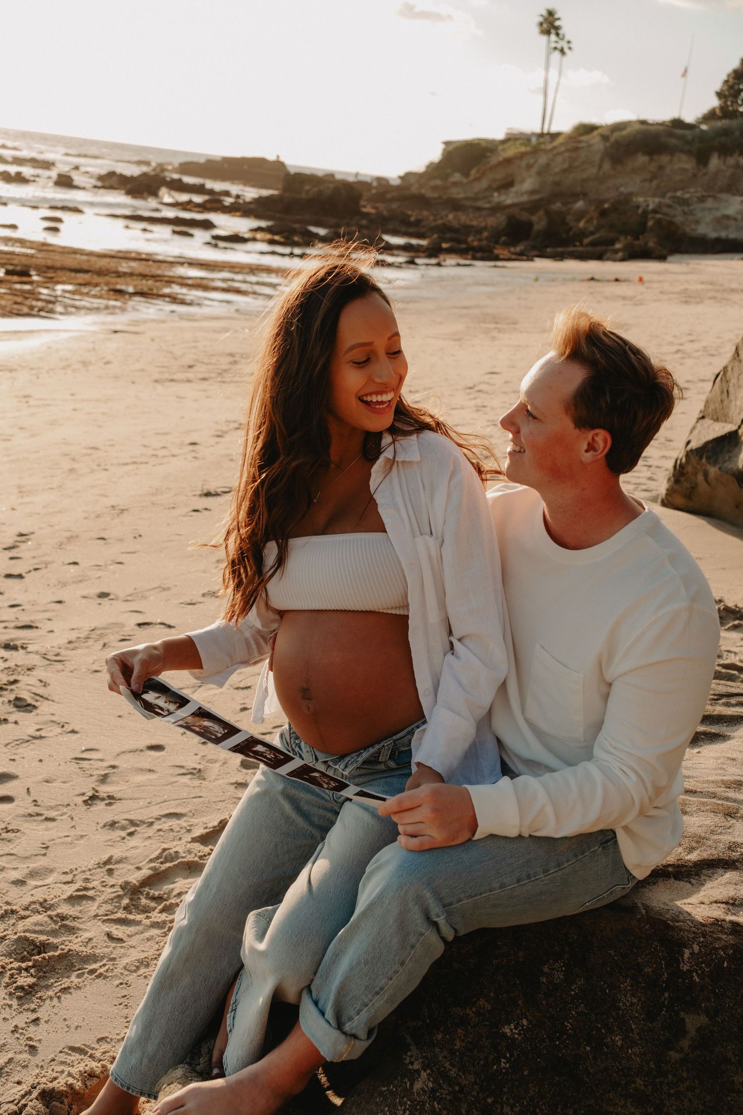 Pregnant couple sitting on beach rocks at sunset, holding ultrasound photo, smiling at each other, wearing white tops and jeans.