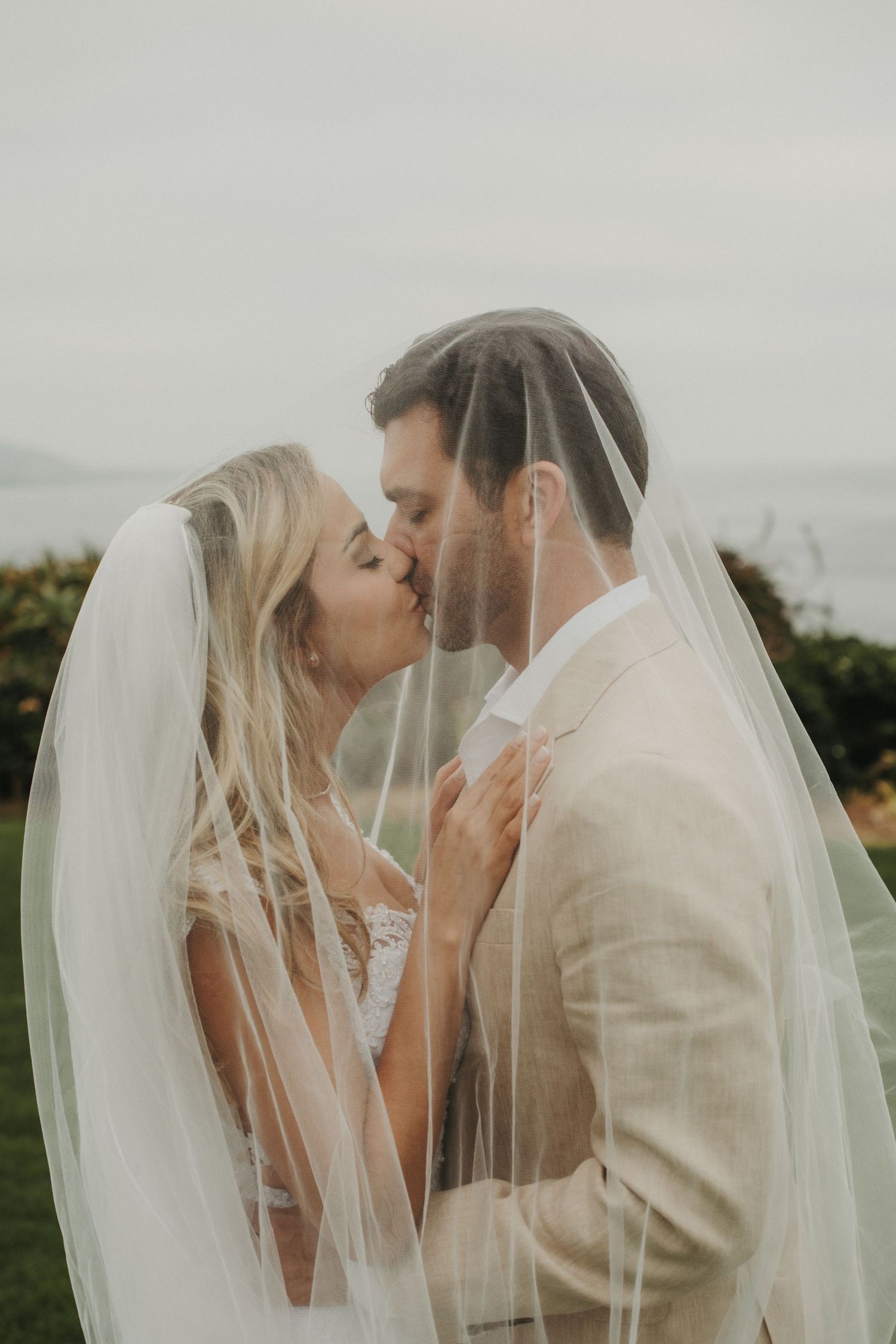 Newlyweds kissing romantically beneath a sheer bridal veil with ocean views behind them.
