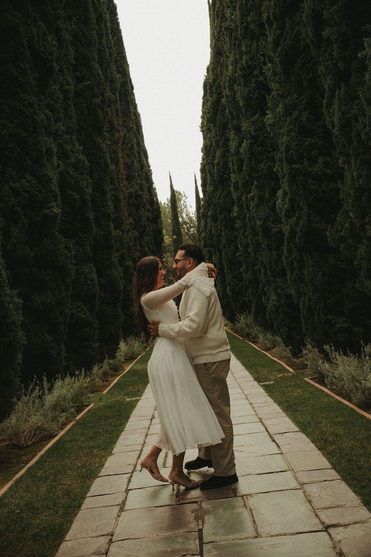 Couple embracing on a stone pathway lined with tall cypress trees in a romantic garden setting.