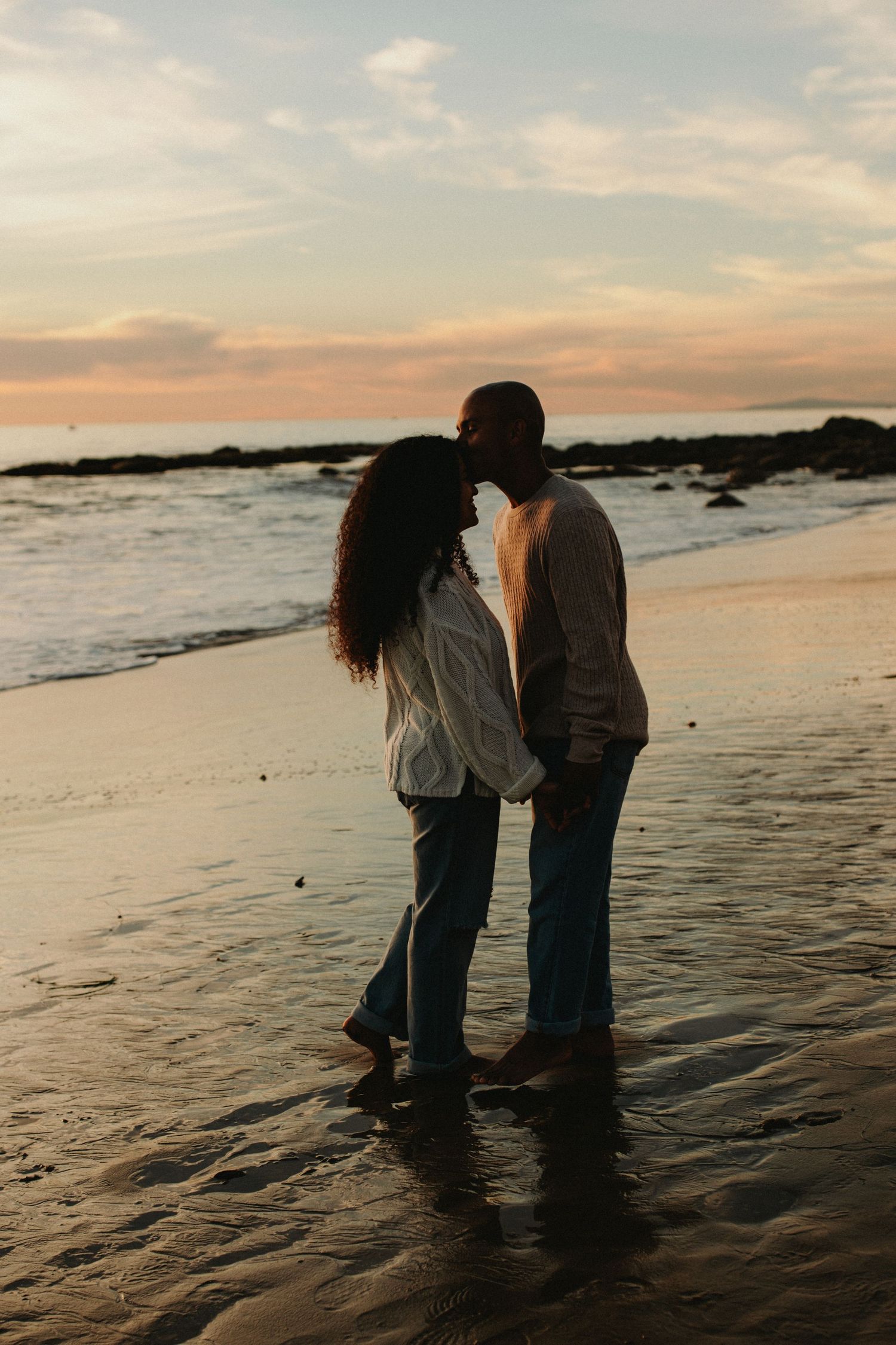 Couple sharing a romantic moment on a sandy beach at sunset, standing barefoot in shallow water with waves behind them.