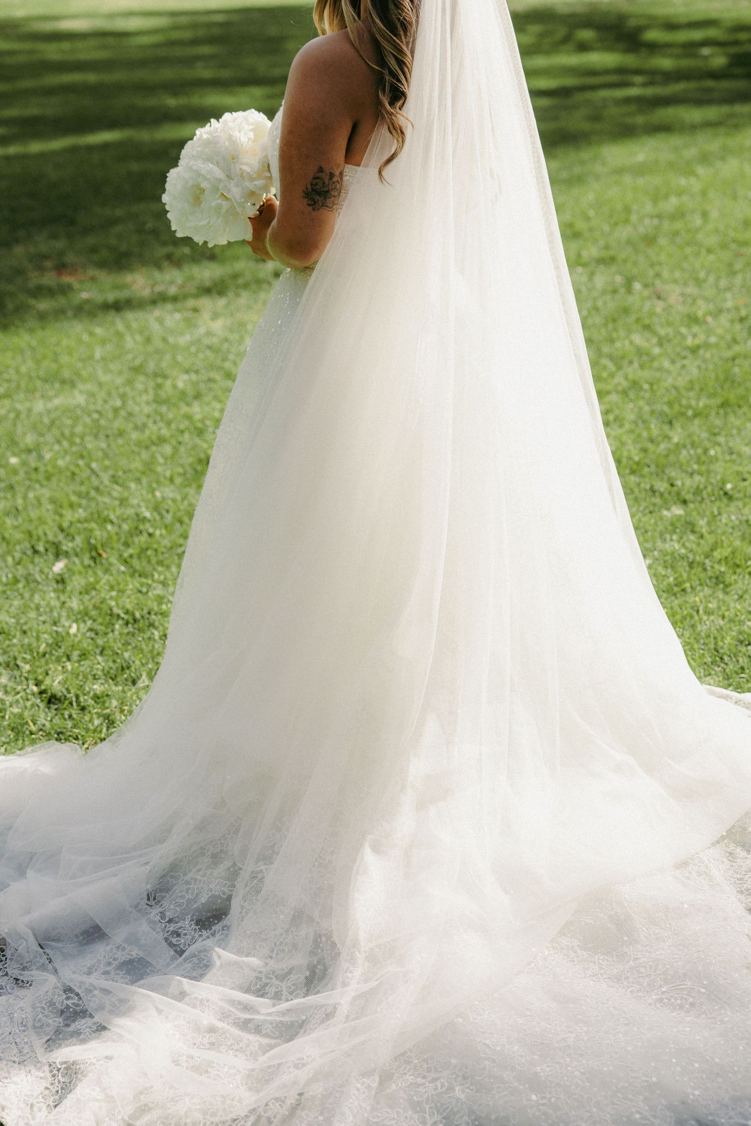 Bride in a flowing white tulle wedding gown with veil and tattoo, holding white bouquet on green lawn.