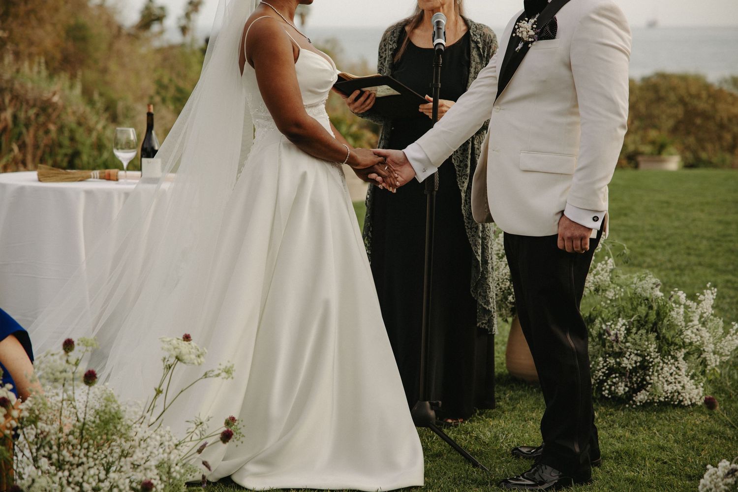 Bride and groom hold hands during outdoor wedding ceremony with officiant in black dress and ocean backdrop.
