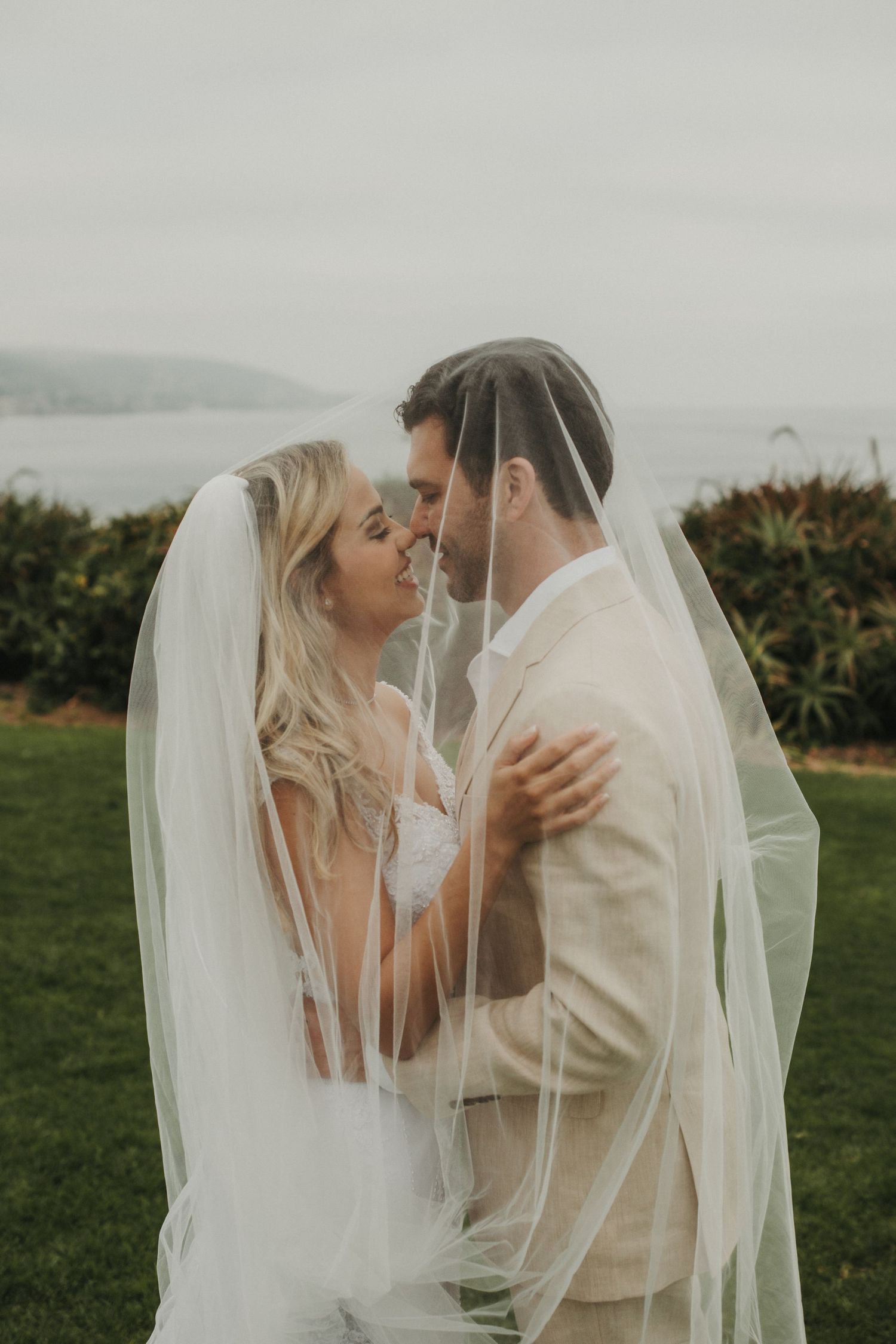 Bride and groom share romantic moment under flowing white veil at outdoor coastal wedding ceremony.