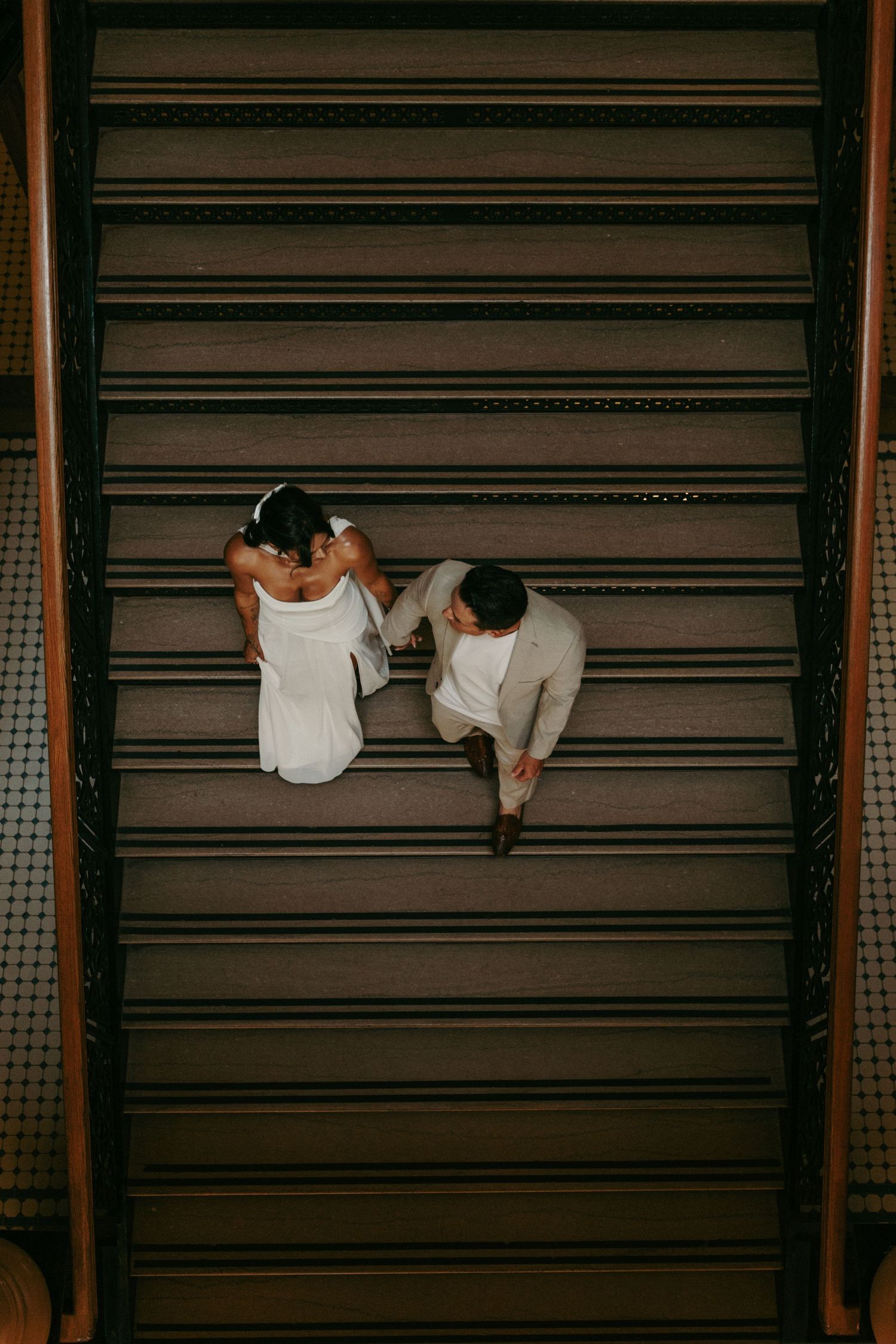 Aerial view of a bride and groom holding hands while walking down elegant wooden stairs on their wedding day.