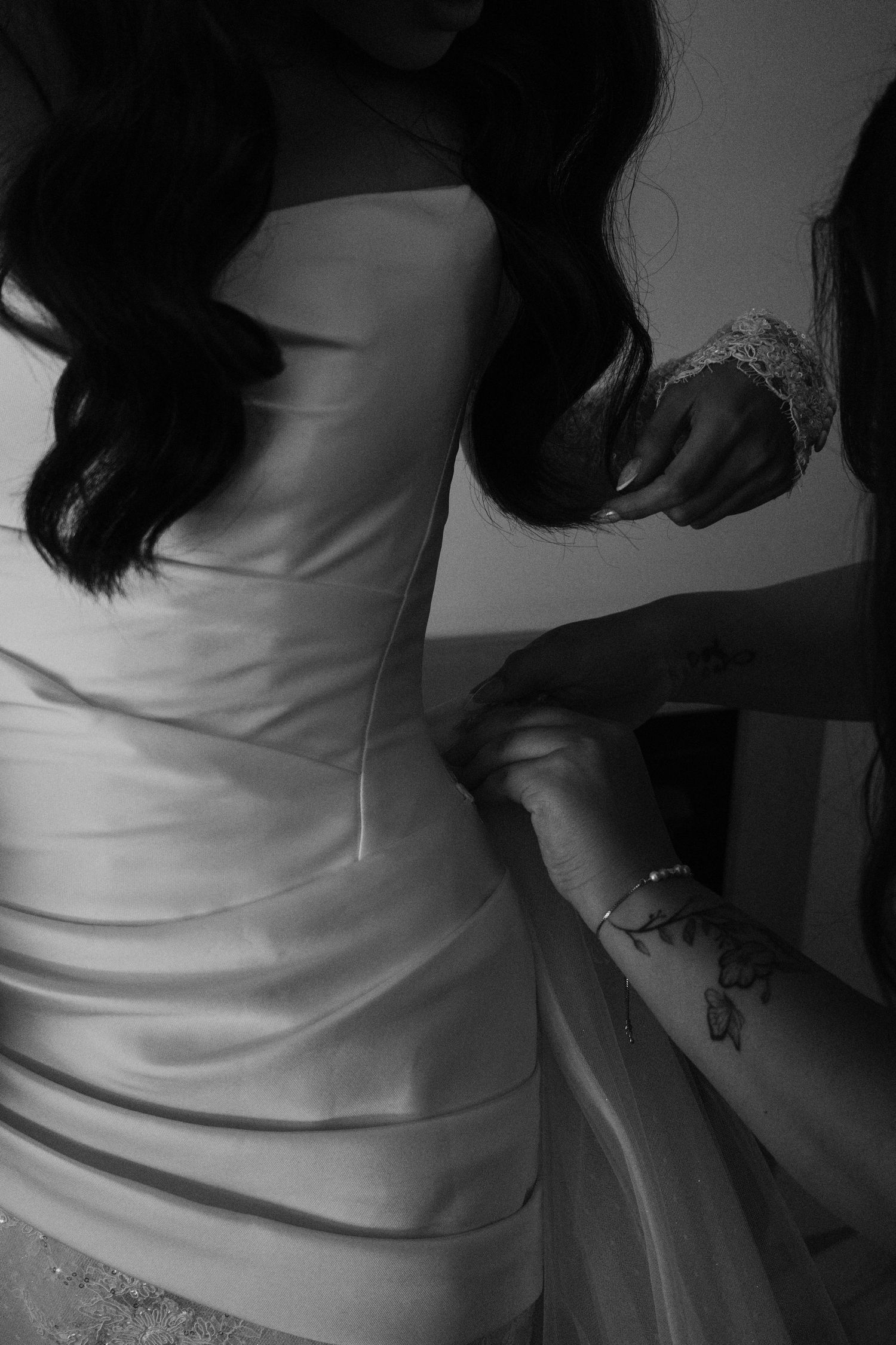 Black and white photo of a bride in a strapless wedding gown being helped by a bridesmaid with a floral tattoo.