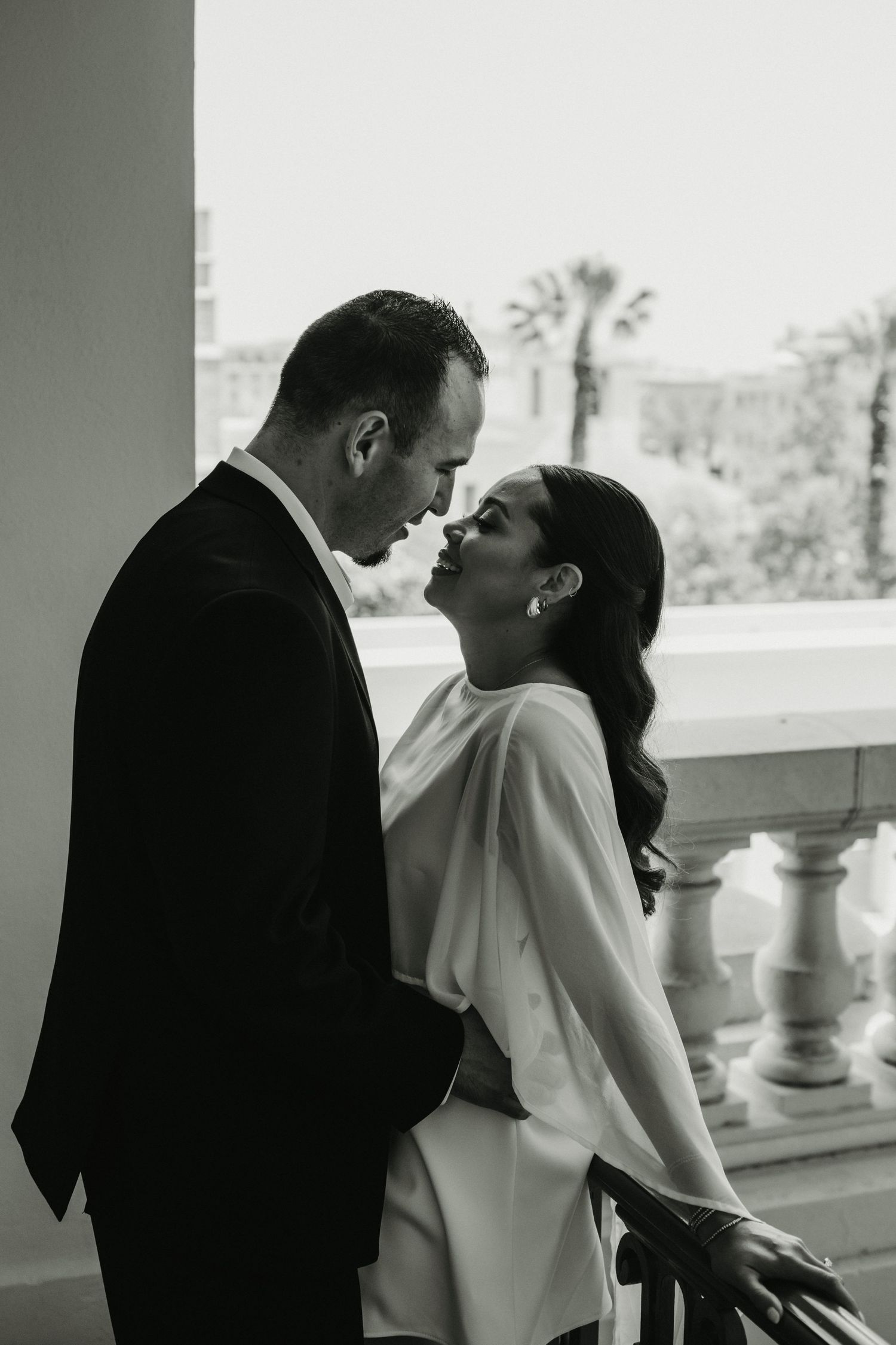 Black and white photo of a couple in wedding attire sharing an intimate moment on a balcony with palm trees in background.