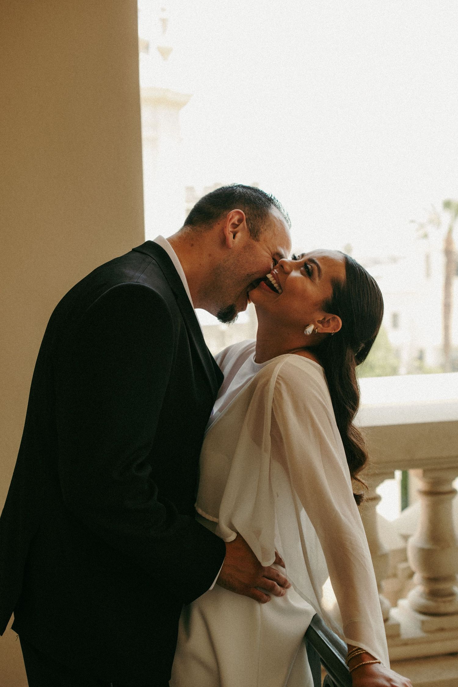 Couple sharing a romantic kiss on a sunlit balcony, groom in black suit and bride in elegant white dress.