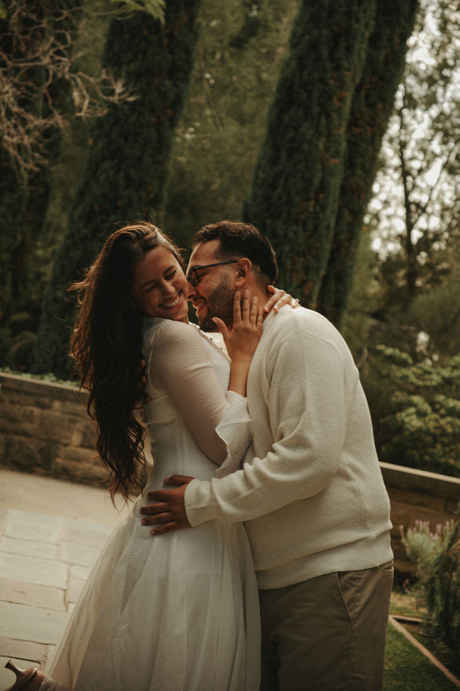 A couple shares a romantic embrace outdoors, the woman in a white dress smiling as the man kisses her cheek.
