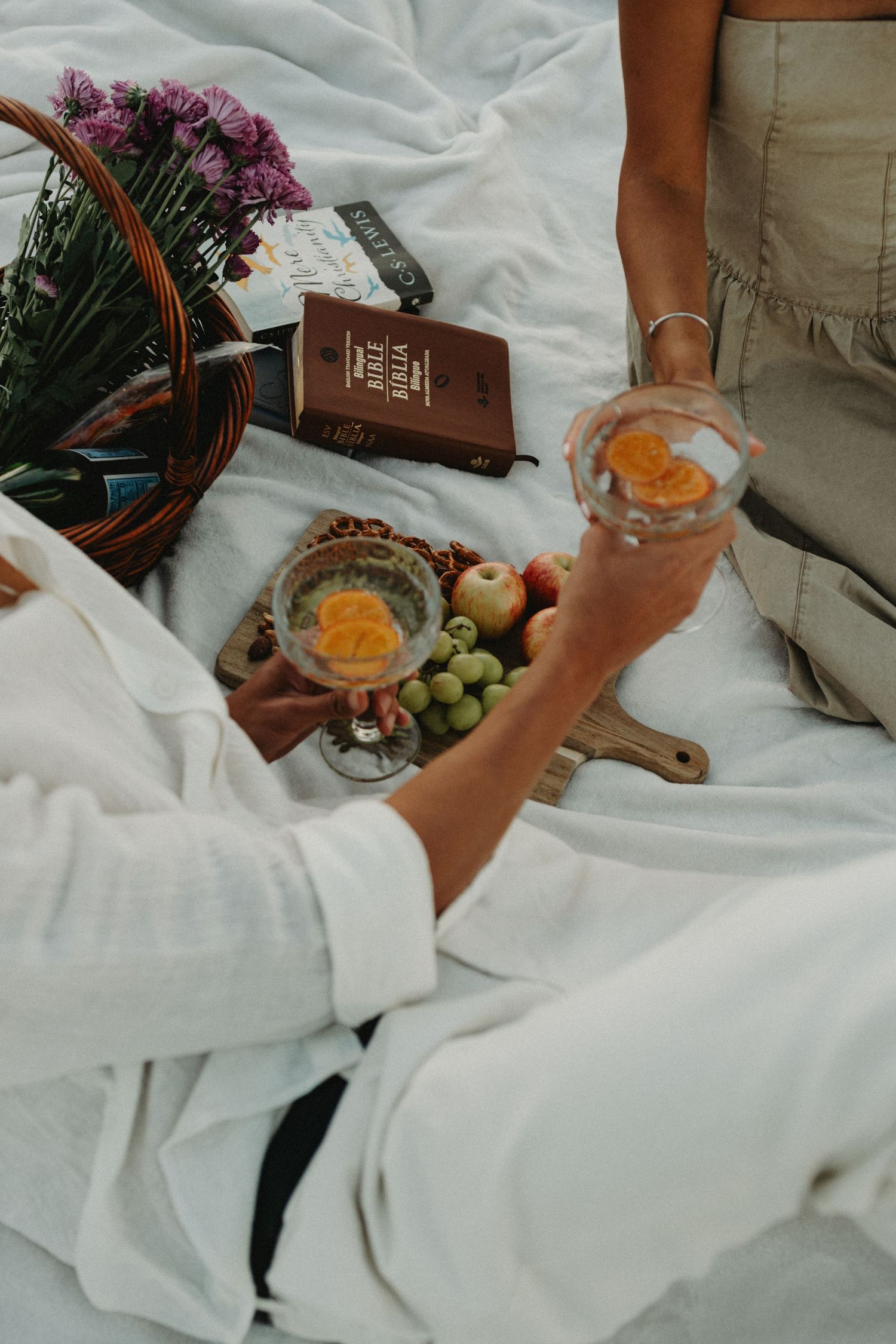 Two women enjoying a picnic with fruit, drinks, and a Bible on a white blanket outdoors.