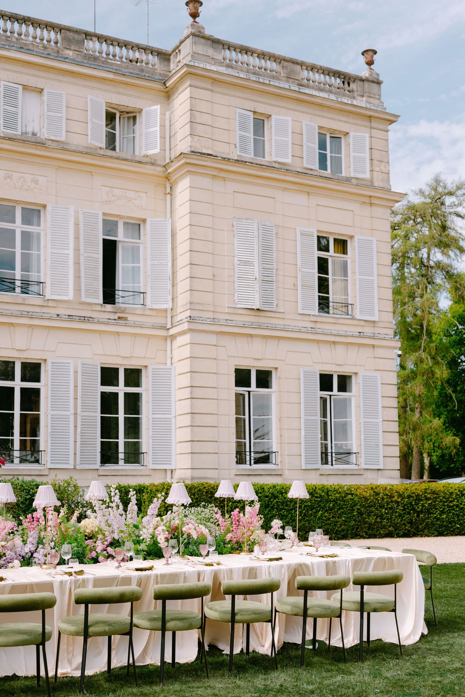 Elegant outdoor wedding reception set on manicured lawn before a grand French château with pink floral centerpieces.