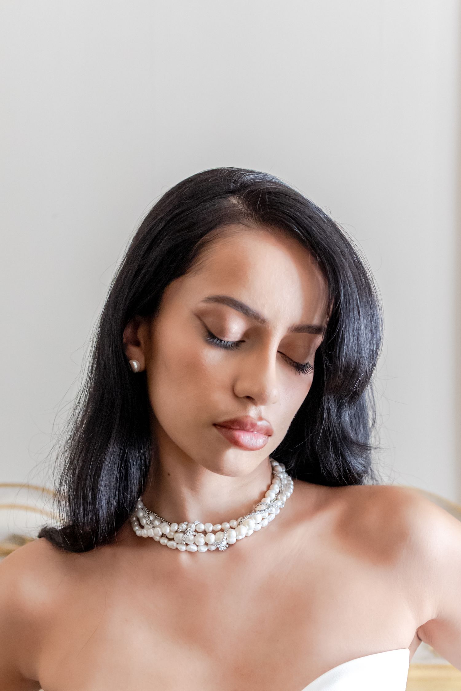 Woman with dark hair wearing a multi-strand pearl necklace and pearl earrings, eyes downcast in elegant bridal style.