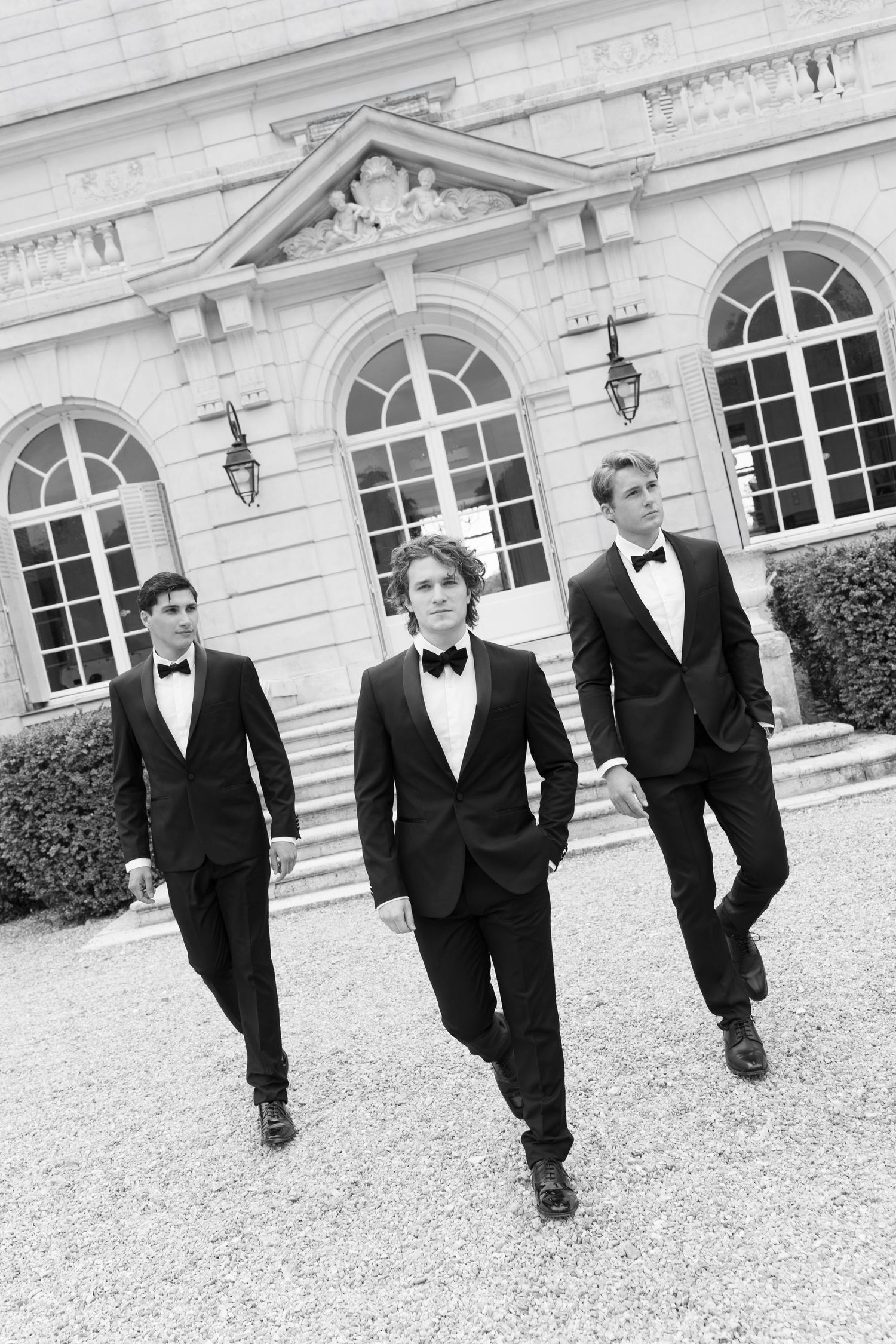 Three young men in black tuxedos and bow ties walk confidently in front of an elegant stone building.