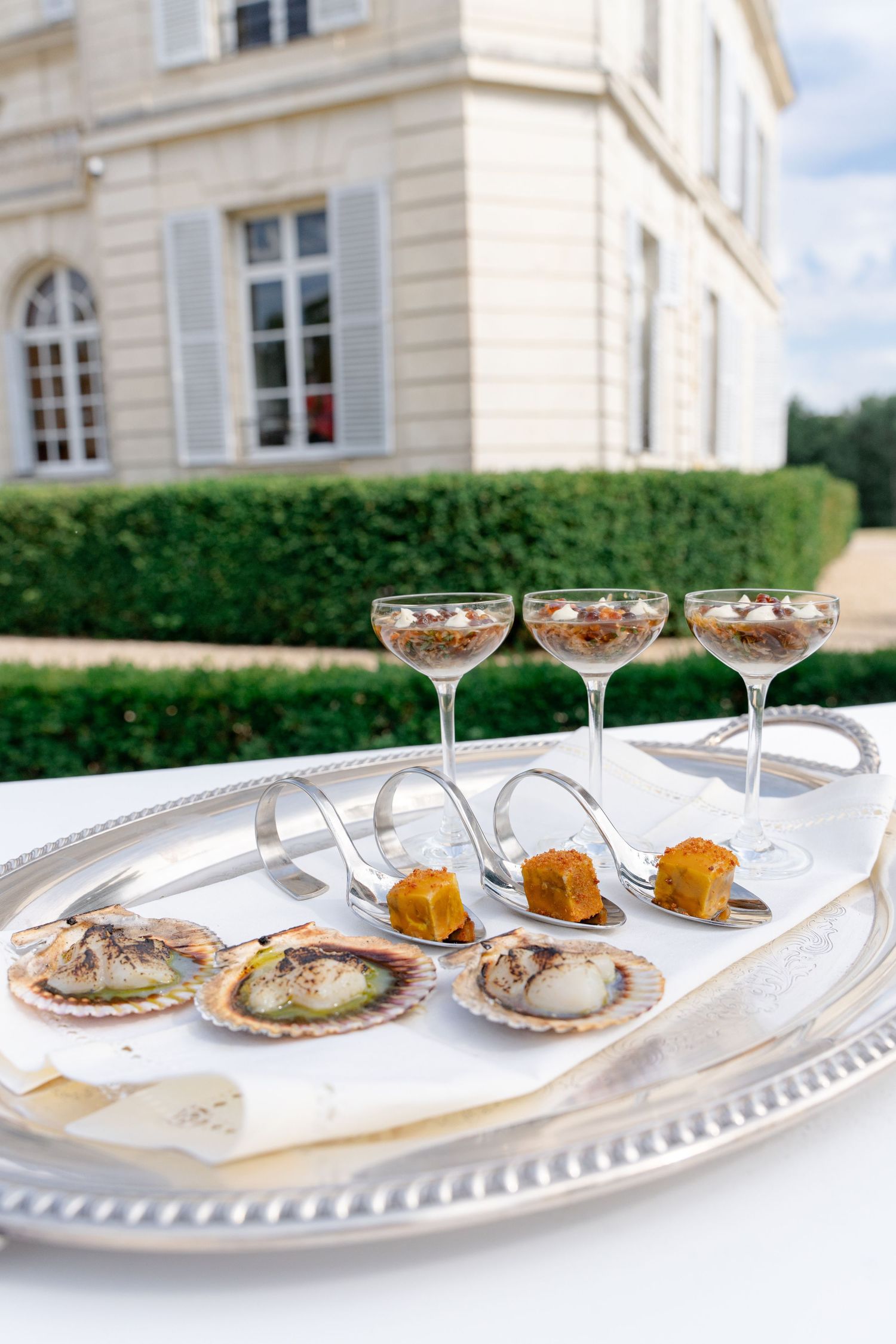 Elegant silver tray with scallops, cocktail glasses, and spoons with orange garnish, served outdoors at a French chateau.