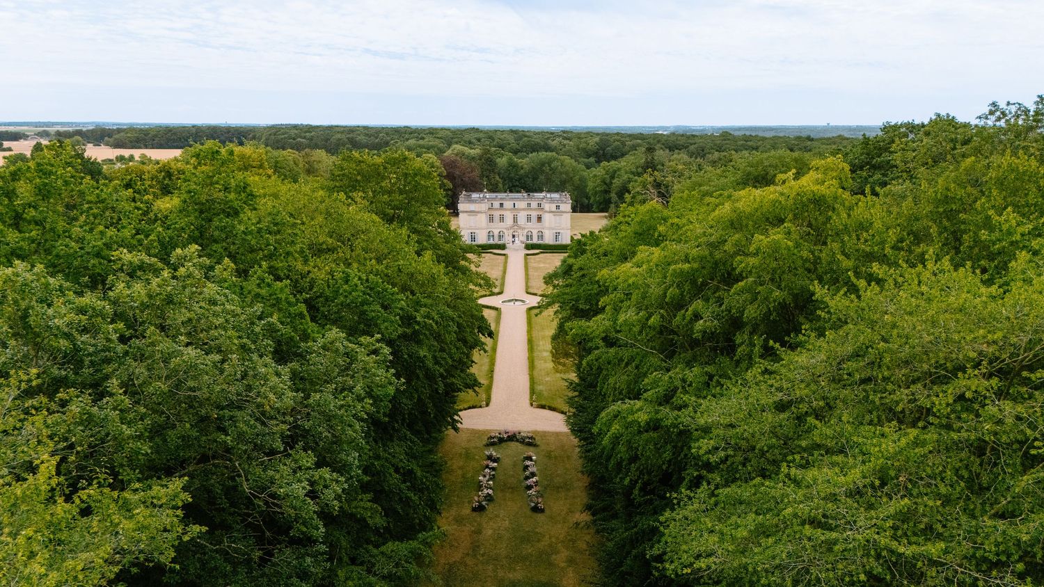 Aerial view of a grand French château framed by lush tree-lined allée leading to formal gardens.