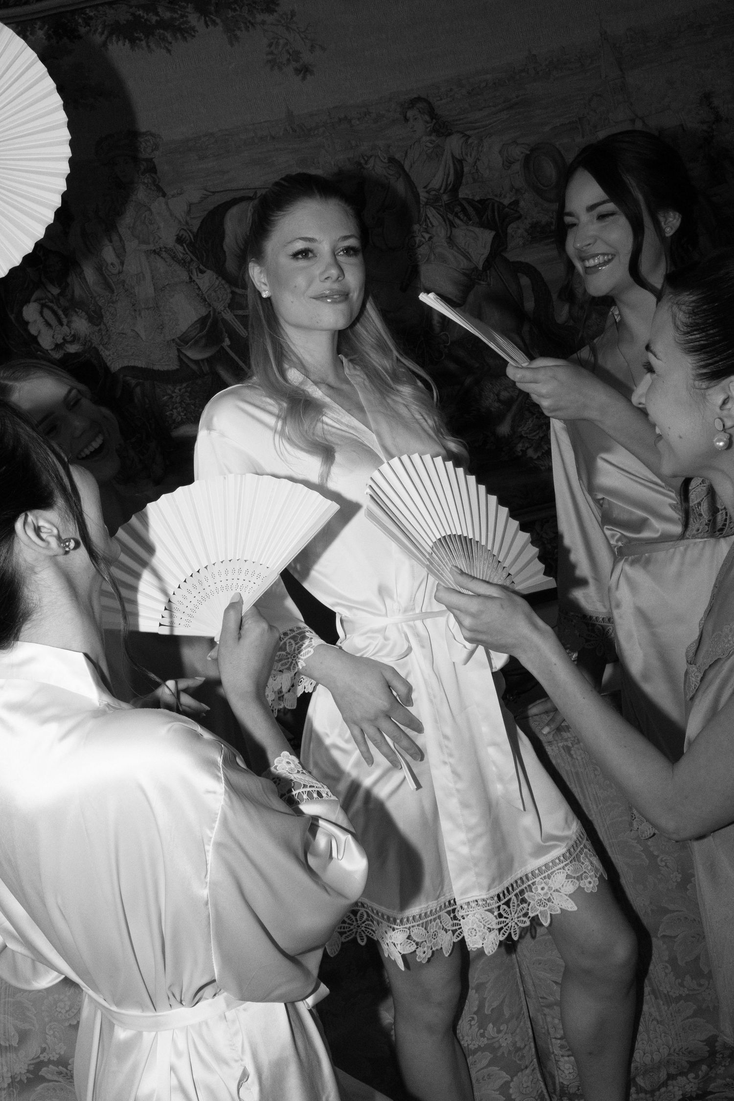 Black and white photo of bridesmaids in robes holding decorative fans surrounding a smiling bride during wedding preparations.