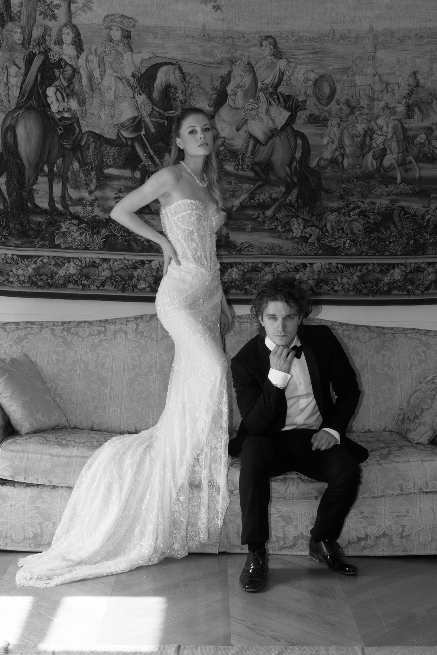 Black and white wedding photo of bride in lace gown standing beside groom in tuxedo on ornate sofa.