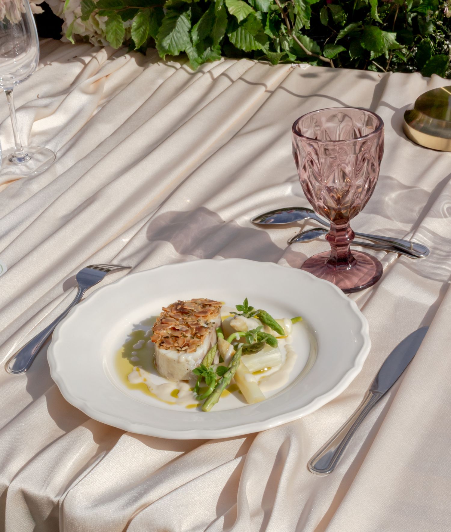Elegant outdoor dining table with a gourmet fish dish, pink goblet, and silverware on a white linen tablecloth.