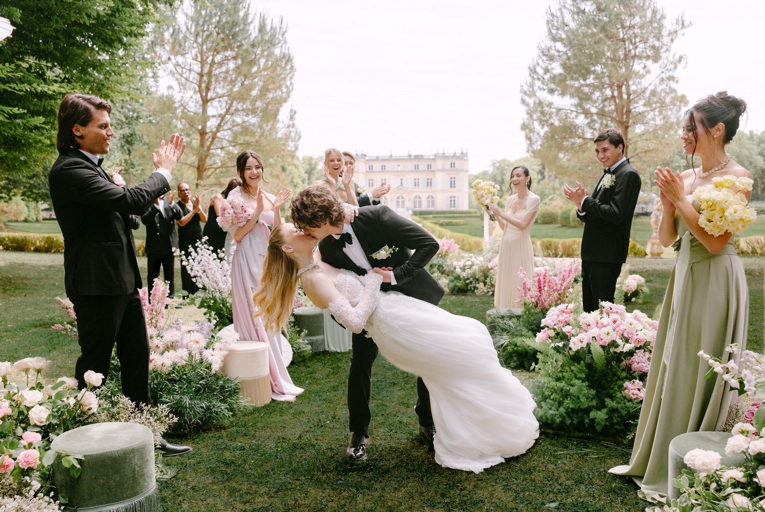 Bride and groom share romantic dip kiss at outdoor wedding ceremony surrounded by floral arrangements and applauding guests.