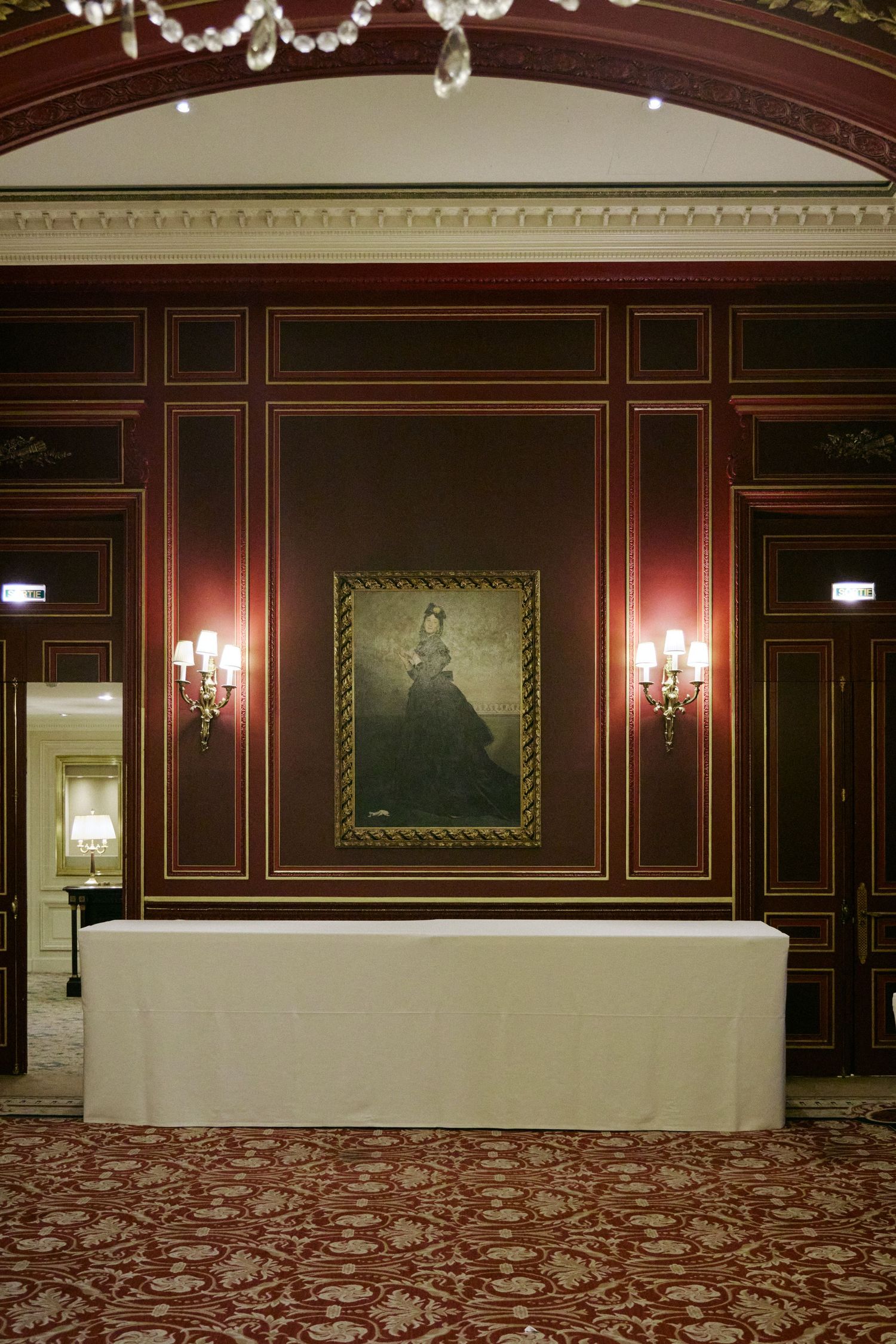 Elegant ballroom with dark wood paneling, gold trim, wall sconces, floral carpet, and a framed portrait above a draped table.