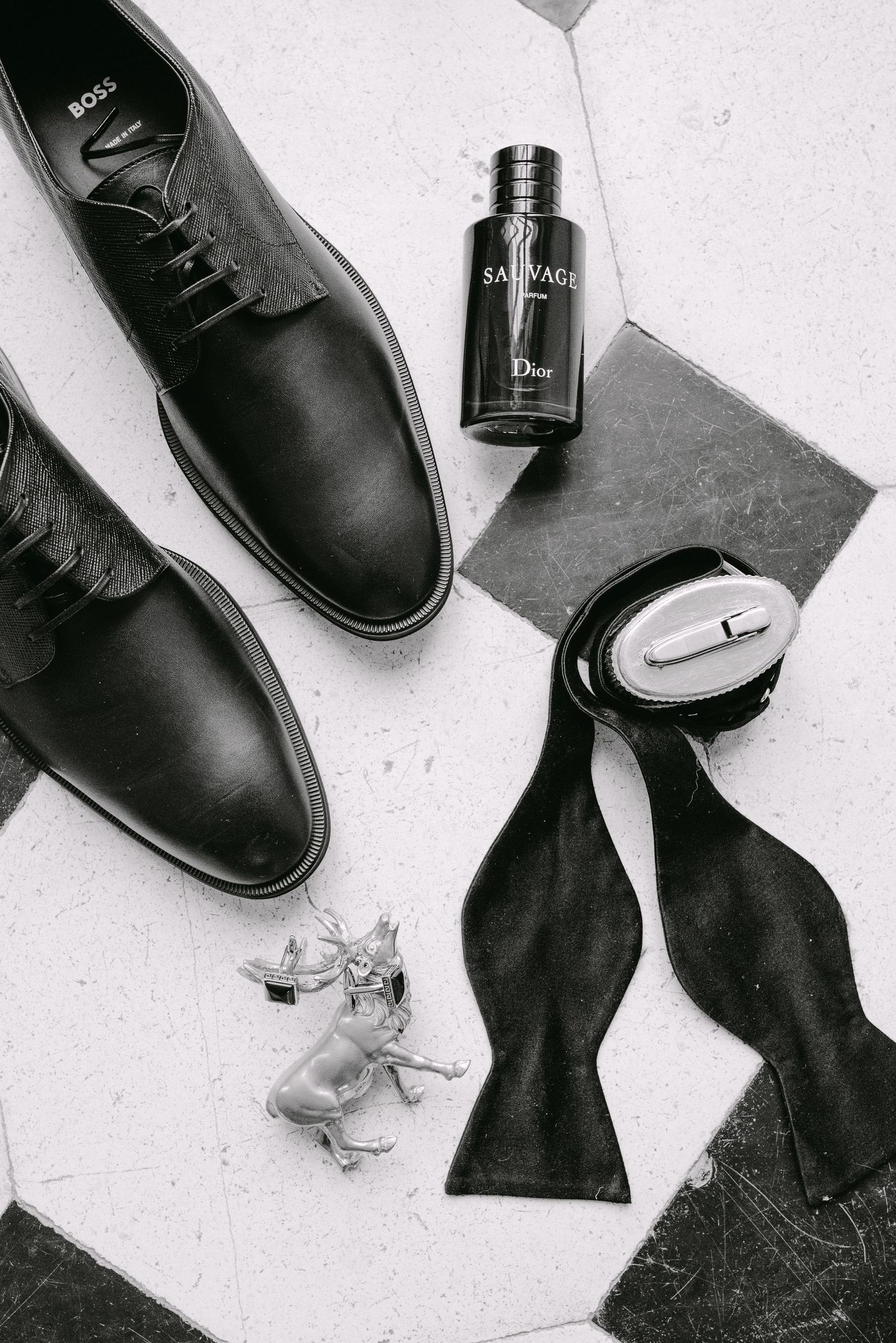 Black and white flatlay of groom accessories including Boss shoes, Dior Sauvage cologne, and bow tie.
