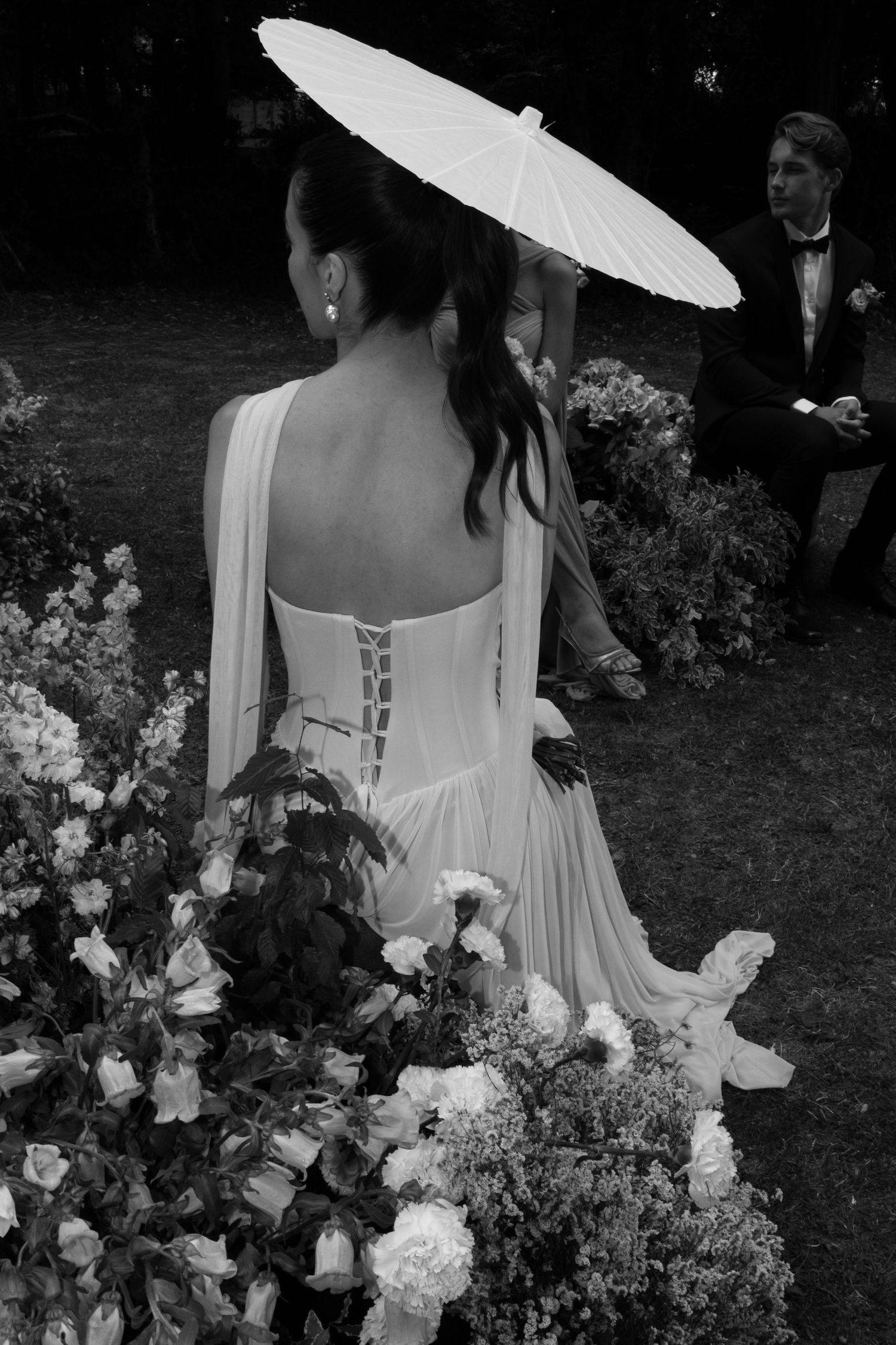 Black and white photo of bride in backless corset gown holding parasol umbrella at outdoor garden wedding ceremony.