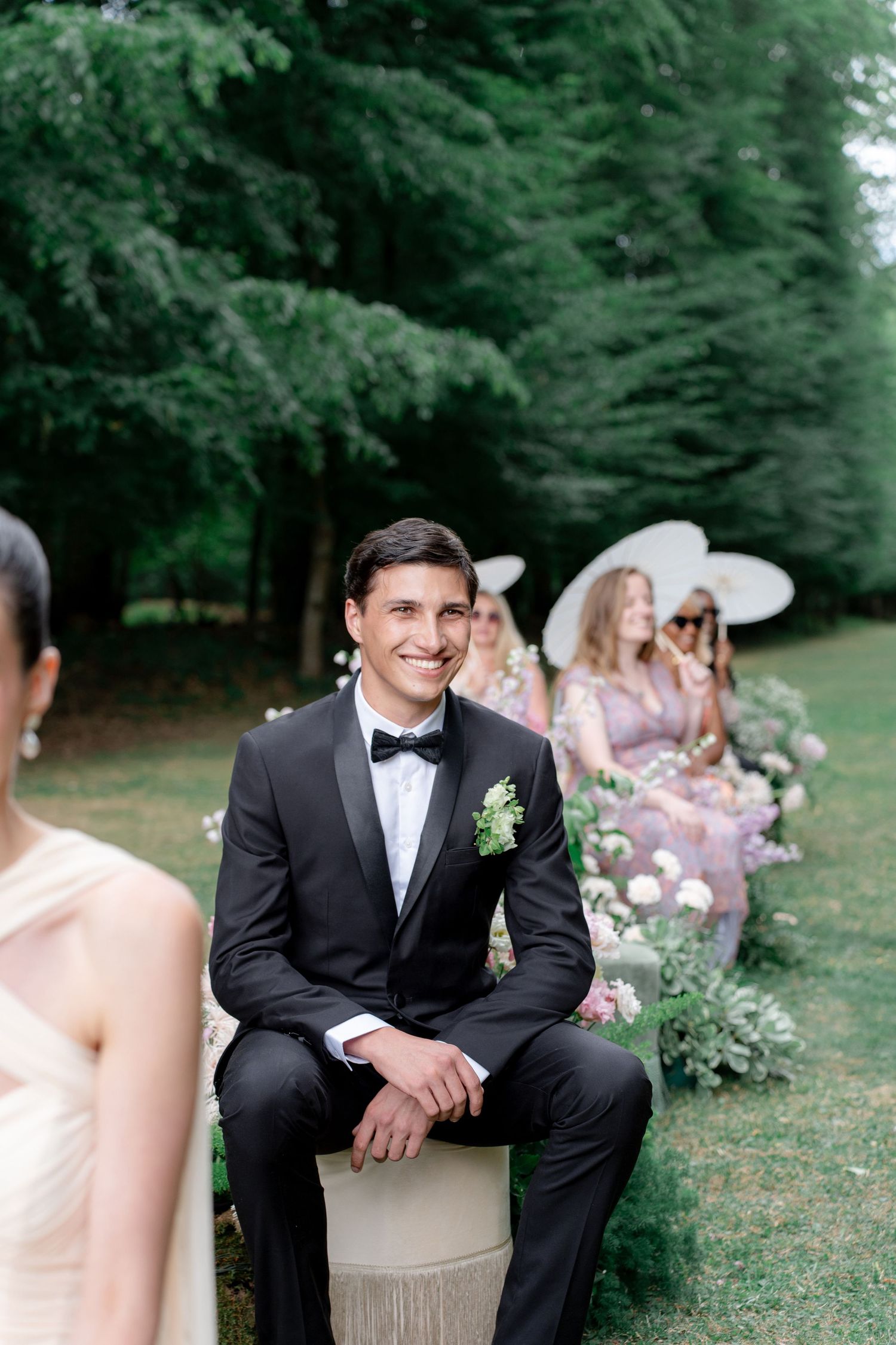 Groom in black tuxedo smiling at outdoor wedding ceremony with bridesmaids in lavender dresses seated behind him.