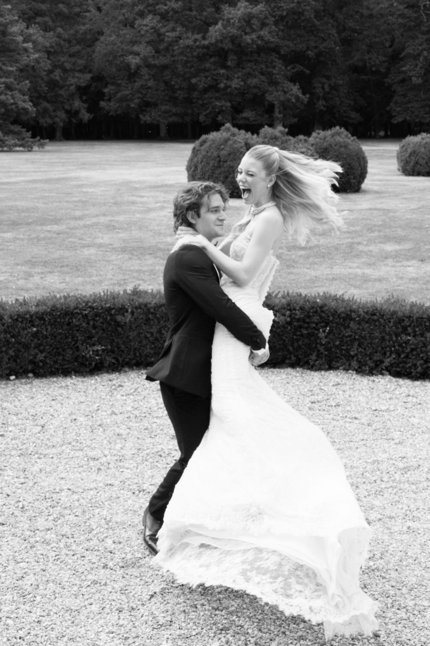 Black and white photo of a joyful bride and groom dancing outdoors on their wedding day in a lush garden.