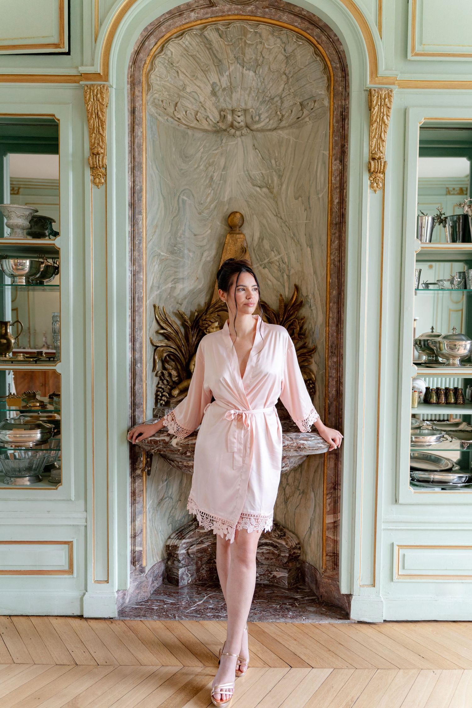 Woman in pink silk robe with lace trim posing in elegant French-style interior with marble alcove.