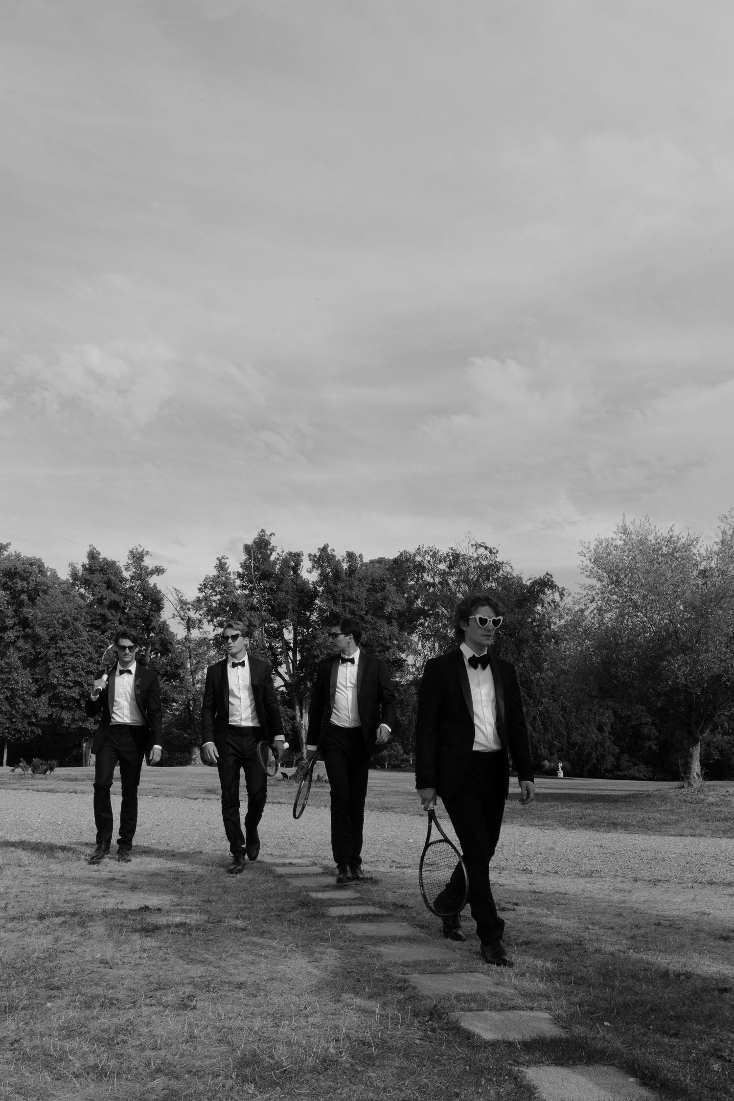 Four men in tuxedos walk through a park, one carrying a tennis racket, in a stylish black and white photo.