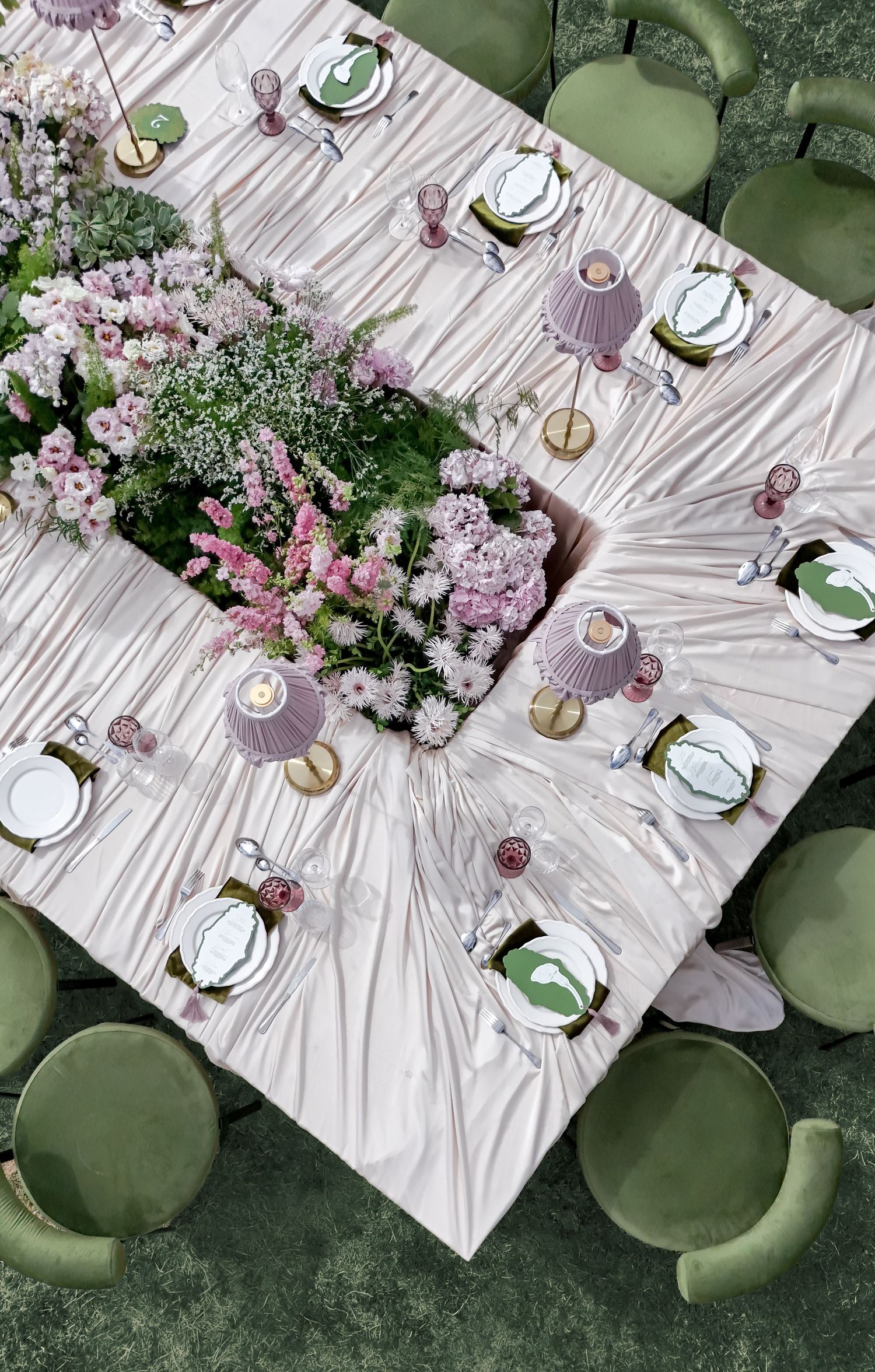 Aerial view of elegant wedding table with pink floral centerpiece, lavender lamp shades, gold accents, and green velvet chairs.