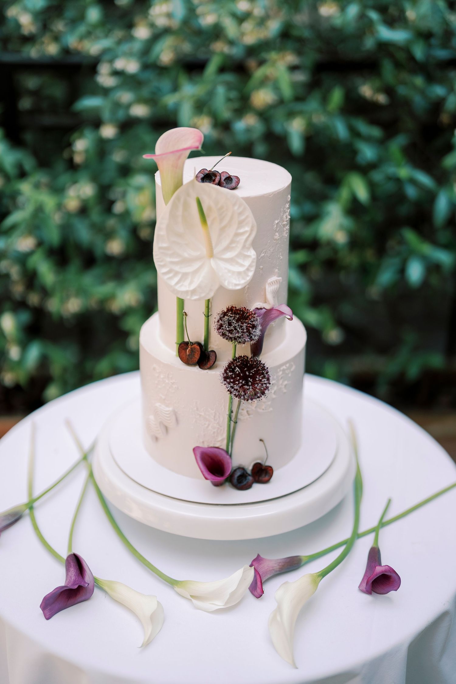 Elegant two-tier white wedding cake adorned with anthurium, calla lilies, and allium flowers on a white pedestal outdoors.