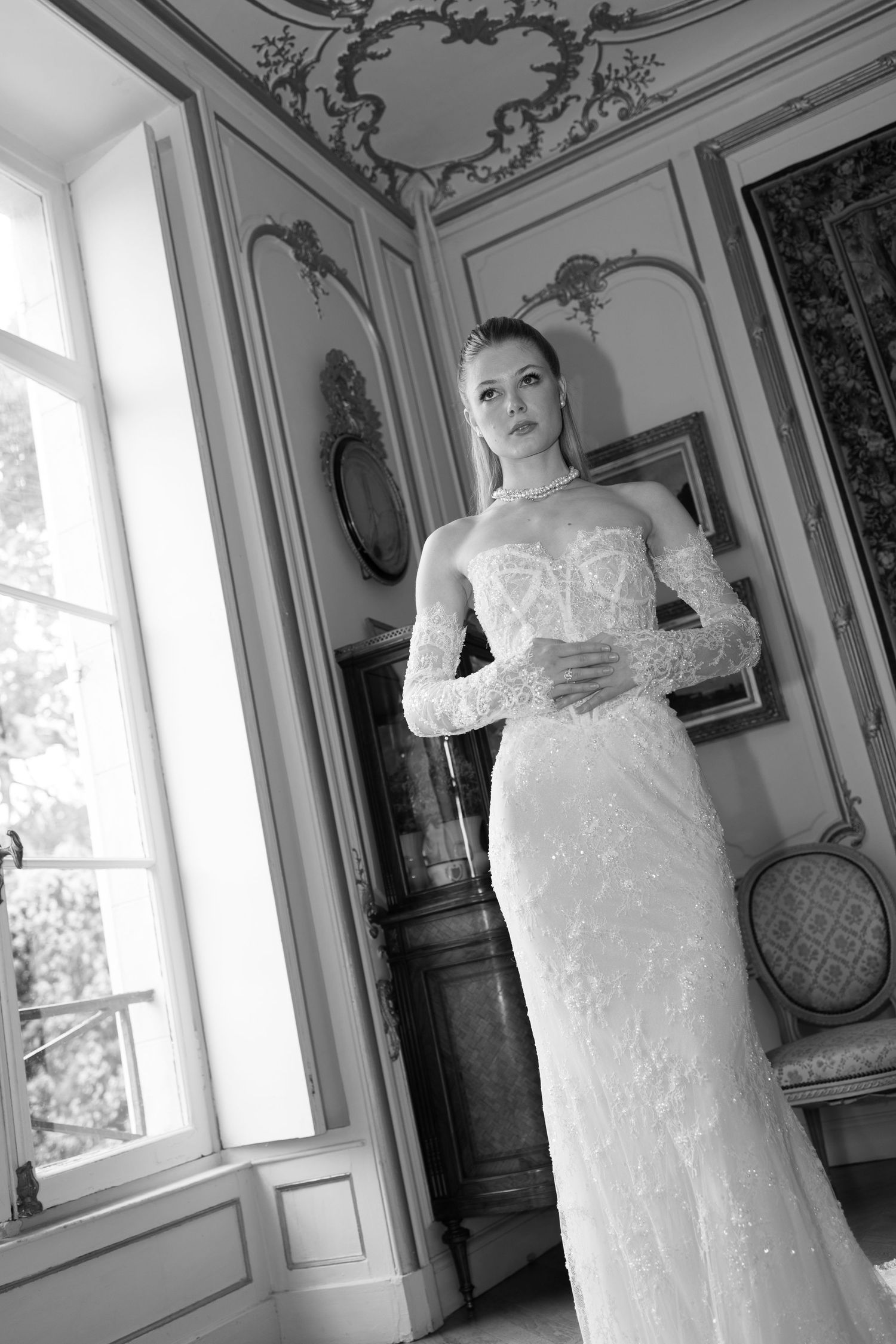 Elegant bride in a lace long-sleeve wedding gown poses in a ornate French-style room, black and white photo.