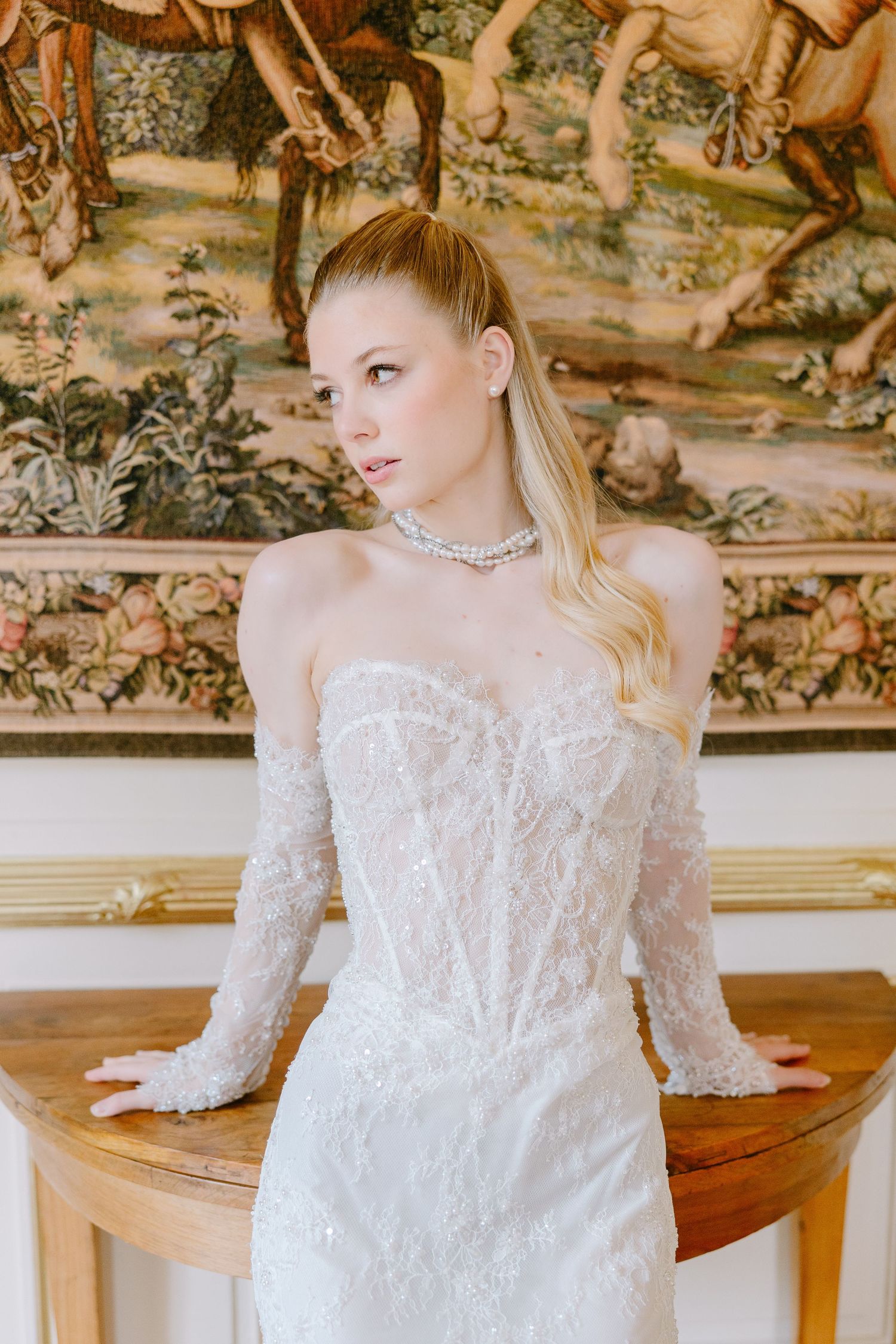 Elegant bride in beaded strapless gown with sheer sleeves and pearl choker, posing before ornate tapestry.