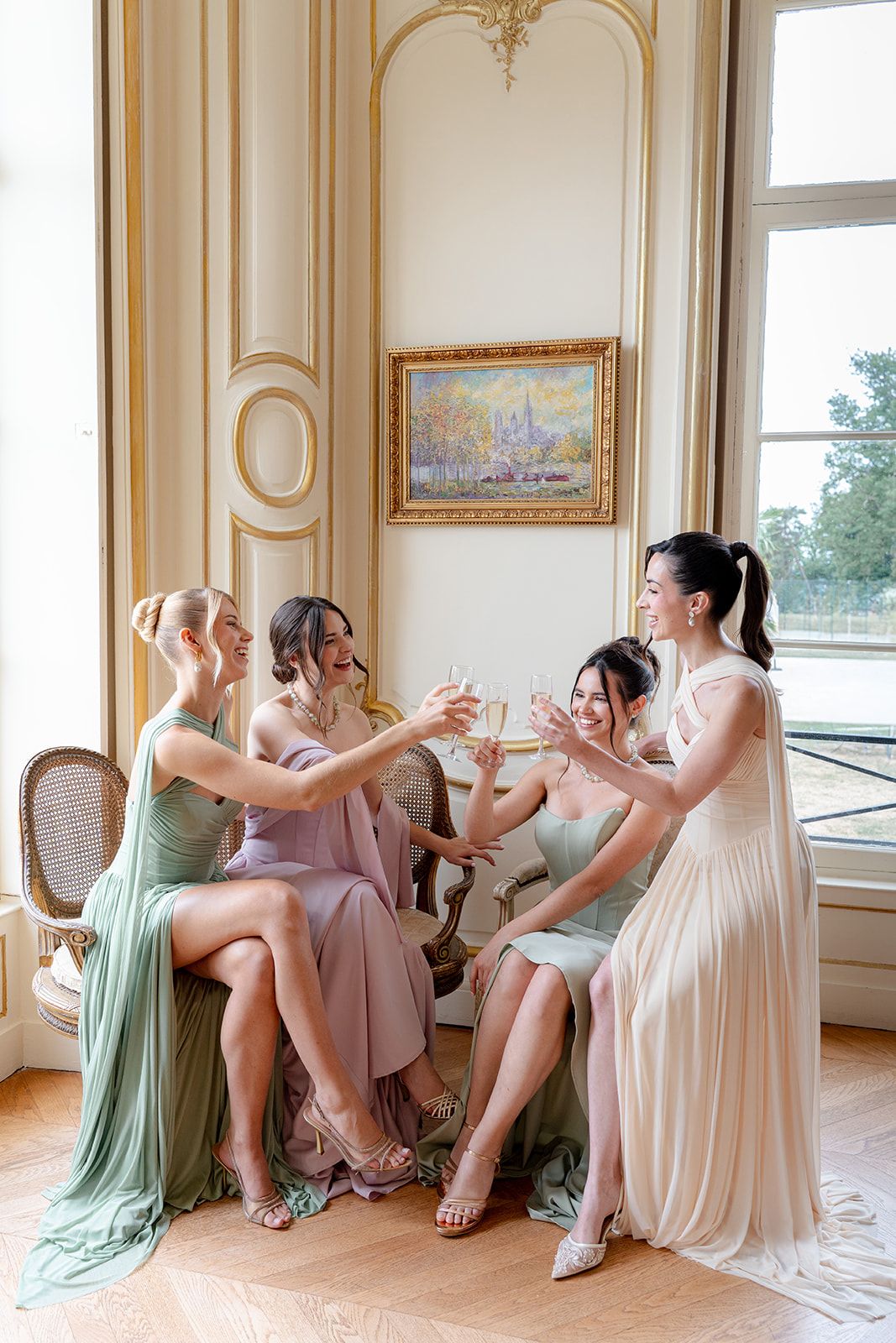 Four bridesmaids in pastel gowns toasting with champagne glasses in an elegant French-style room.
