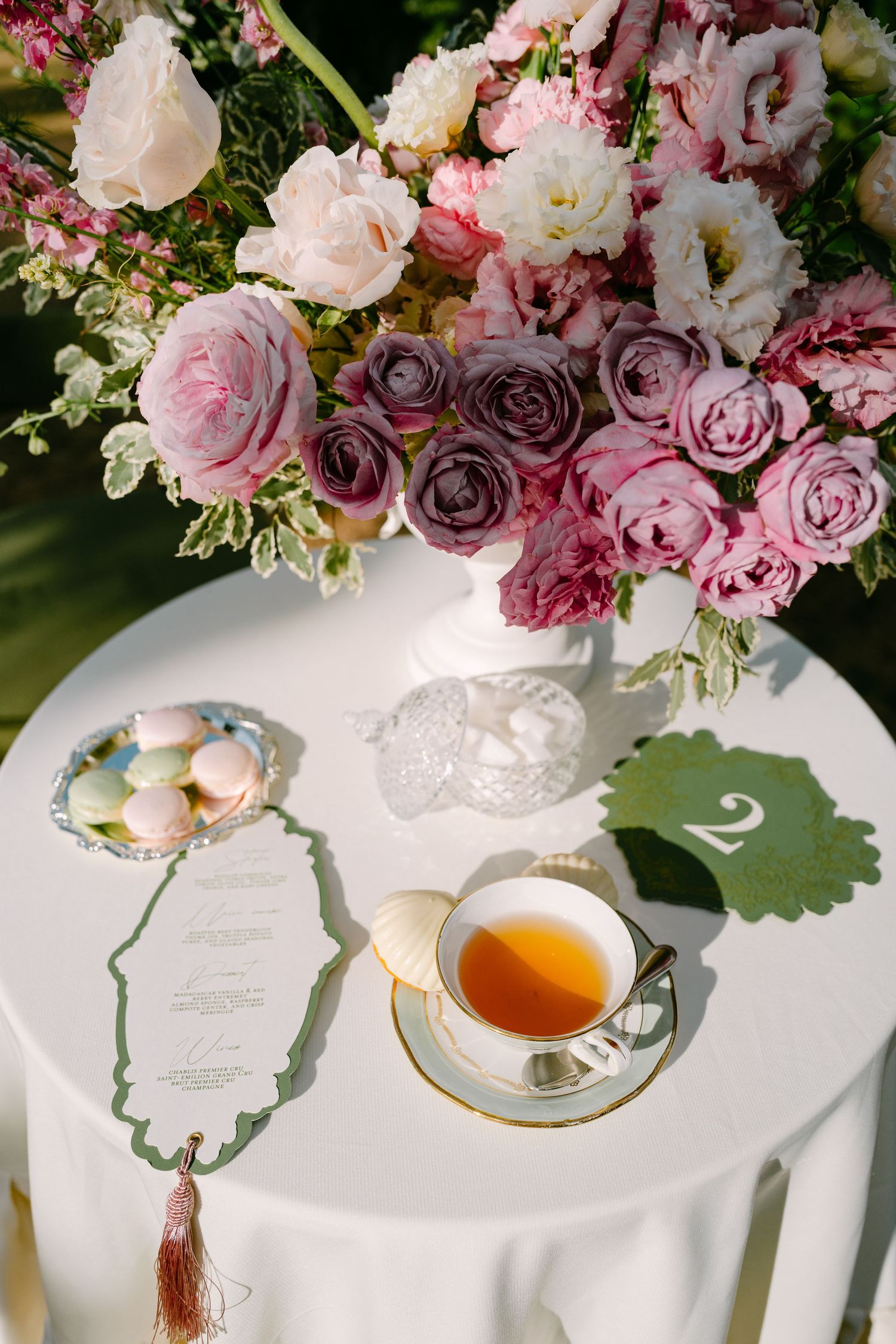 Elegant wedding table with pink roses, tea cup, macarons, and decorative menu card outdoors.