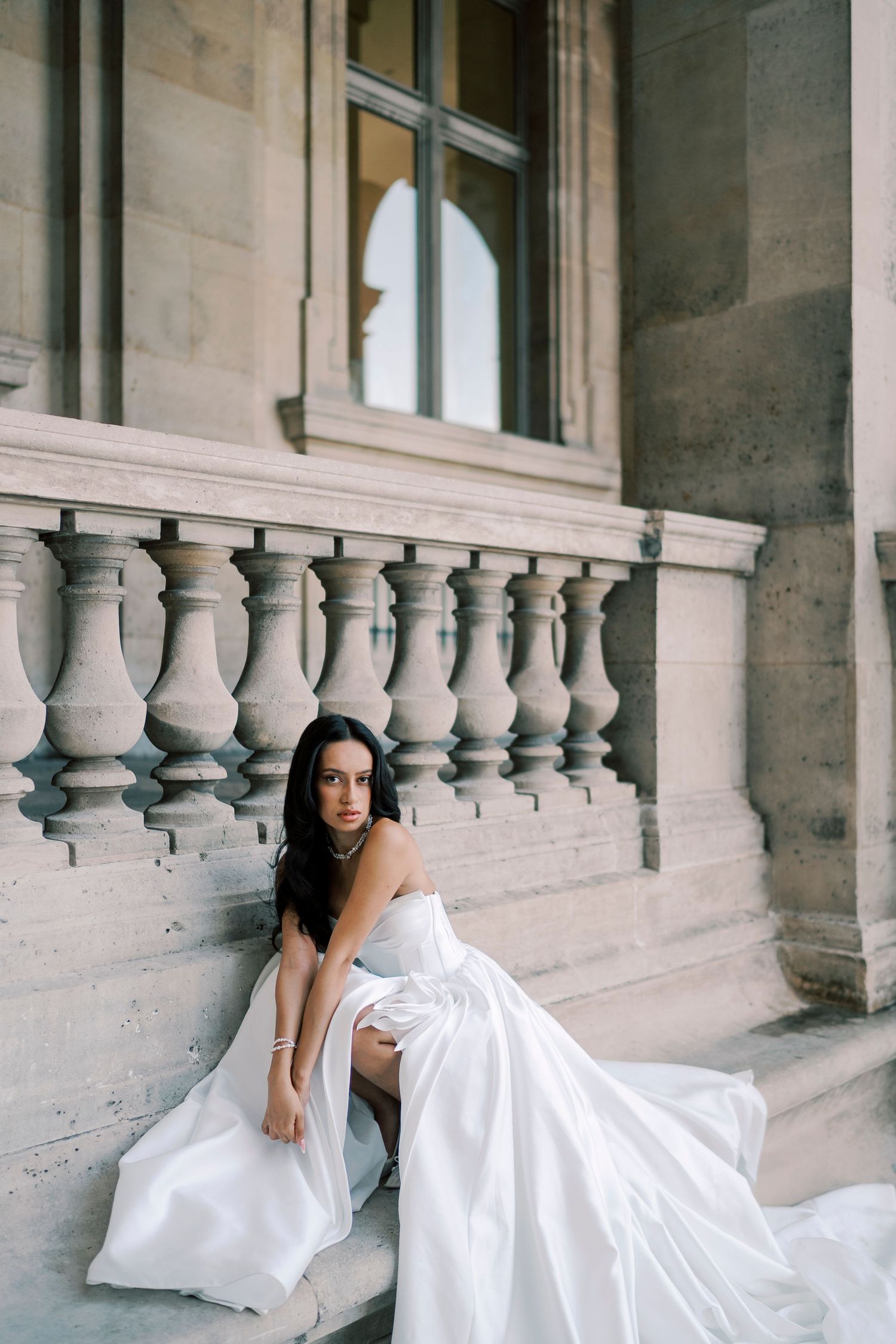 Elegant bride in a flowing white gown sitting on stone steps of a classical building with ornate balustrade.