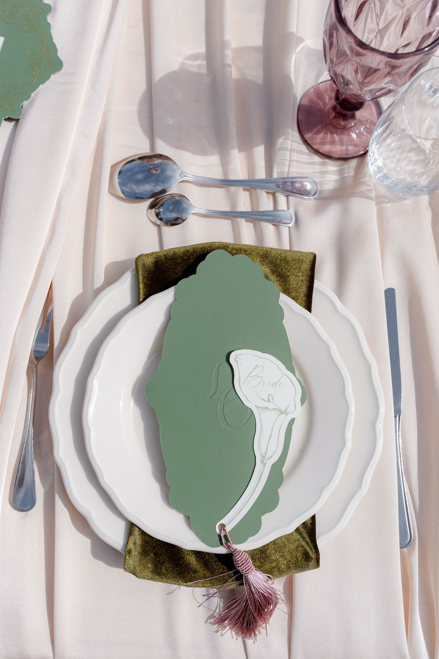 Elegant table setting with white plates, green velvet napkin, sage menu card, silver cutlery, and purple glassware on cream linen.