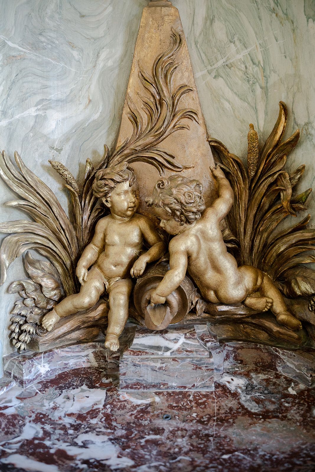 Golden cherub sculptures resting against an ornate marble backdrop with decorative foliage in classical Baroque style.