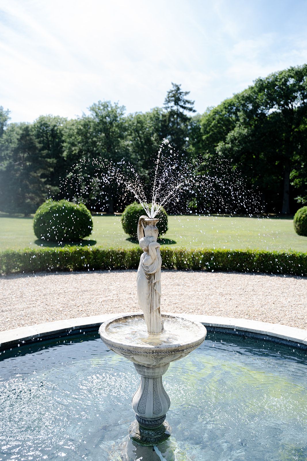Classical stone fountain with water spraying from a figurine statue, set in a formal garden with manicured hedges and trees.