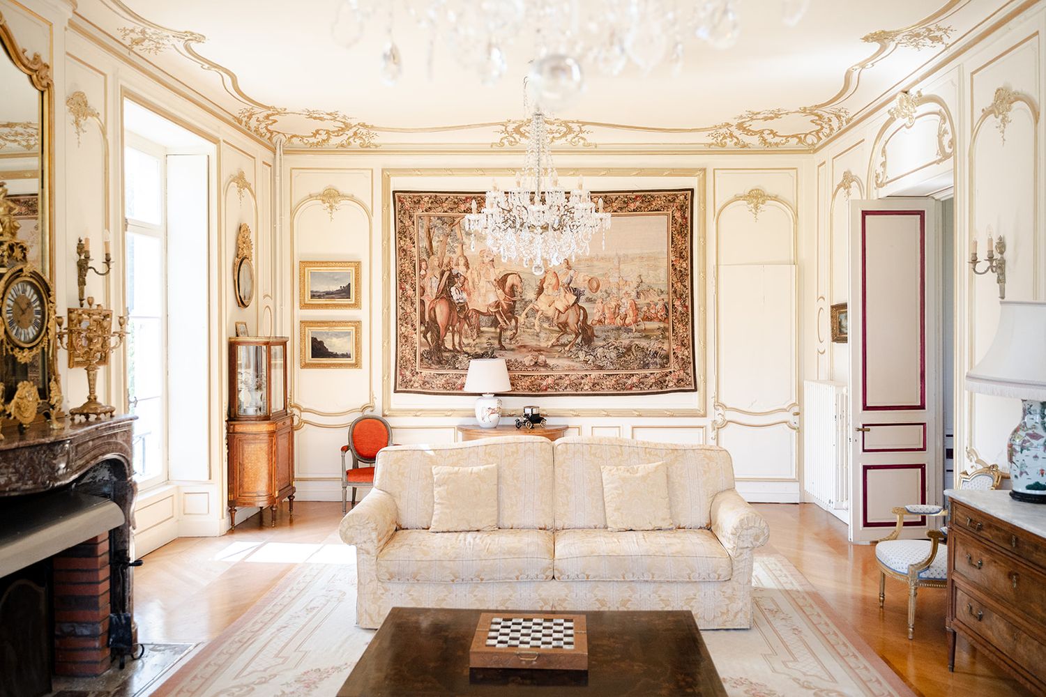 Elegant French-style living room with ornate gold moldings, crystal chandelier, white sofa, and large tapestry wall art.