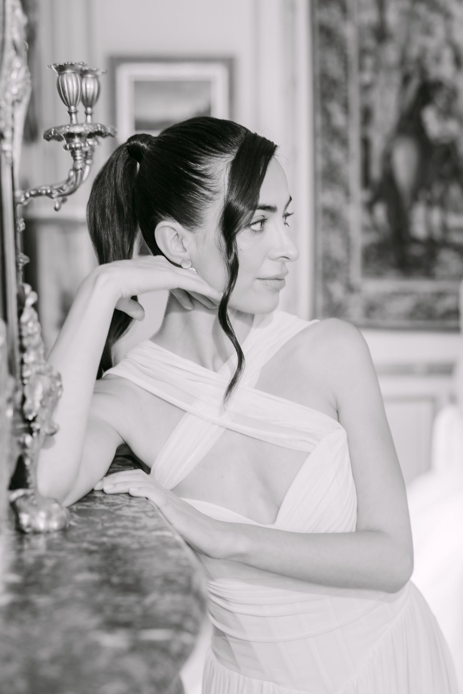 Black and white portrait of a bride in a white halter gown leaning against a marble surface in an ornate room.