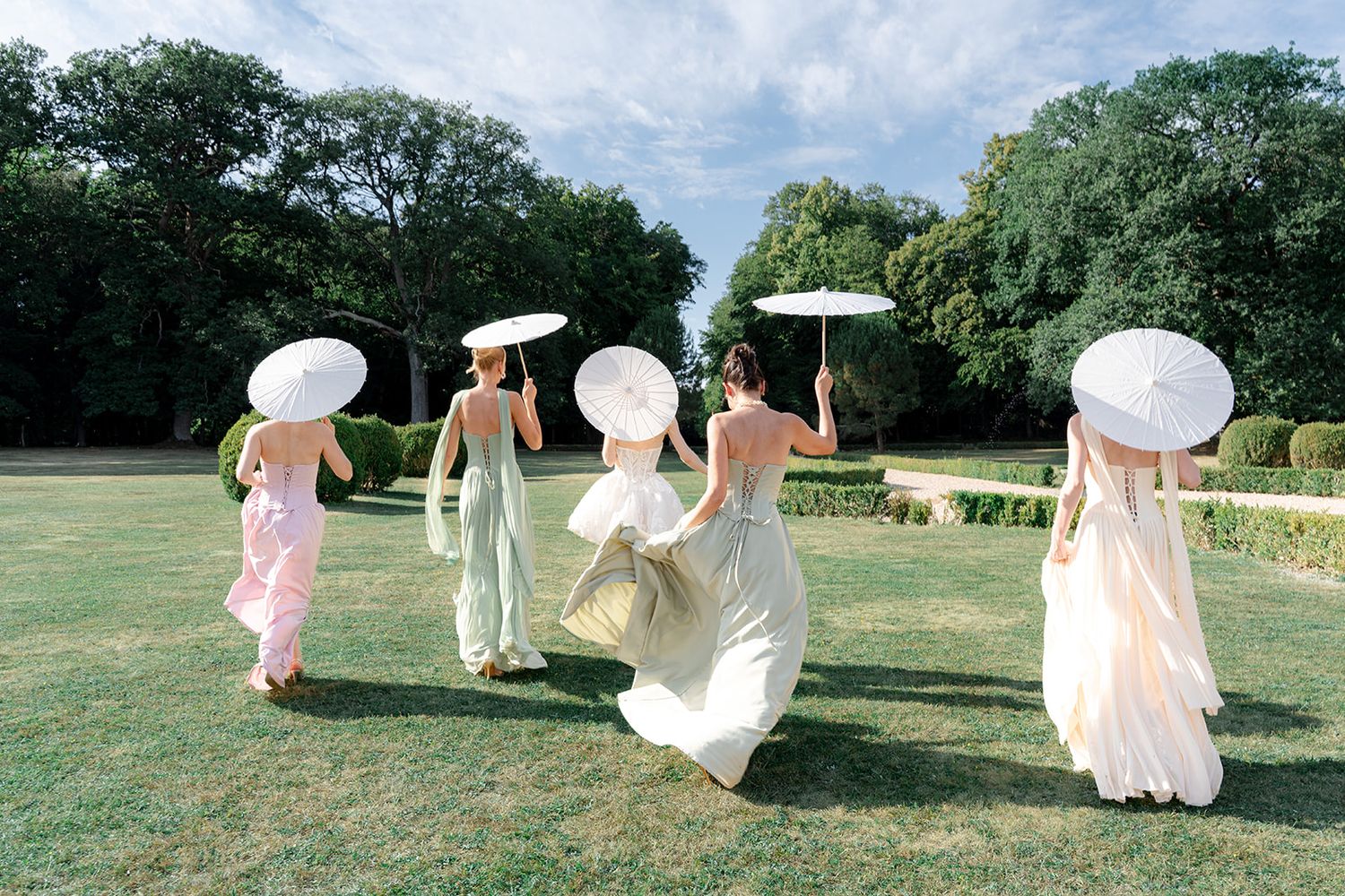 Four bridesmaids in pastel gowns carry white parasols while walking across a lush green garden lawn.